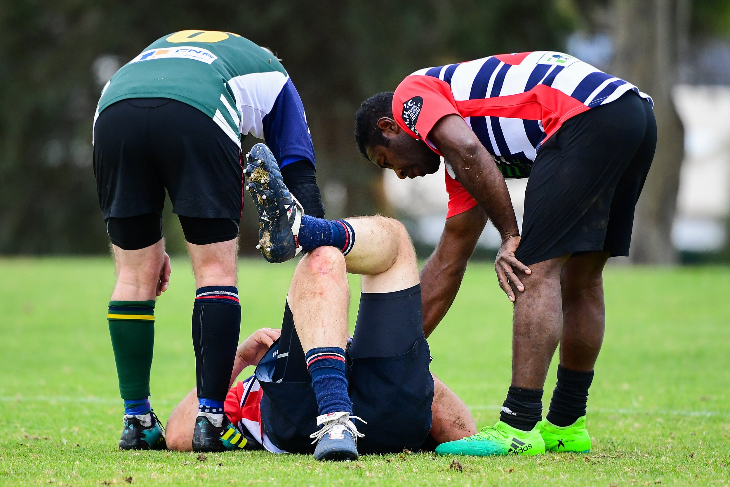 Two rugby players assisting an injured teammate lying on the ground during a match, with one player supporting his leg and the other kneeling beside him.