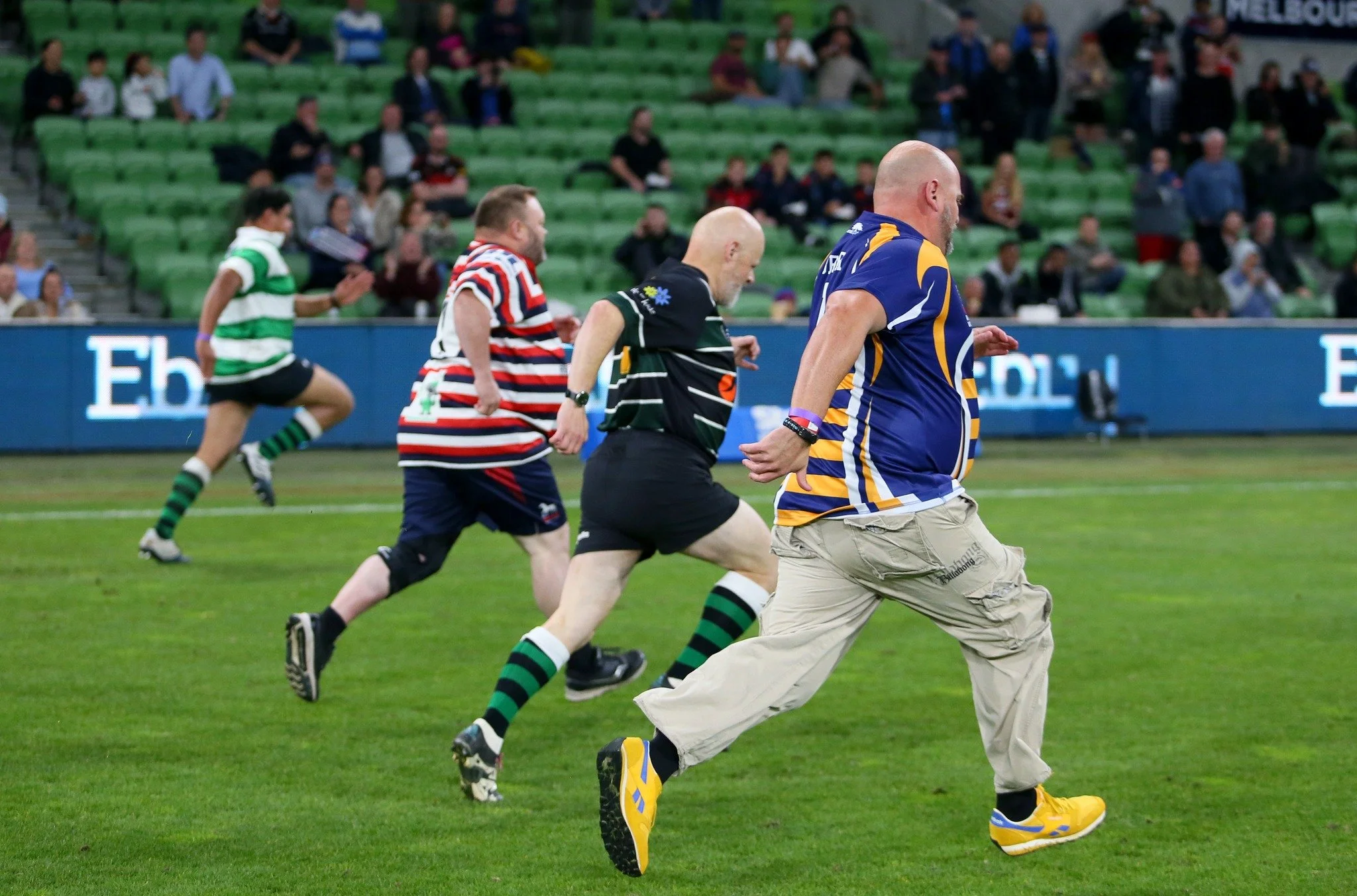 Men in casual sportswear running on a rugby field with green grass, with spectators seated in a stadium in the background.