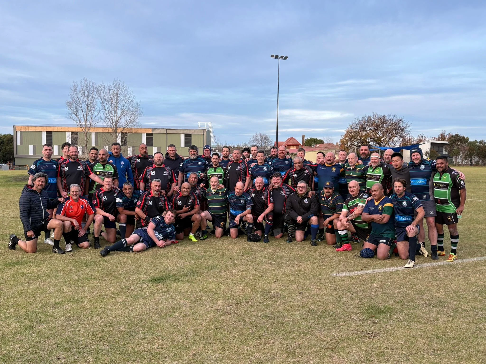 Group of rugby players and coaches posing for a team photo on a rugby field, with some standing and others kneeling or lying in front. The players wear uniforms in various colors including black, navy blue, green, and striped patterns. The background