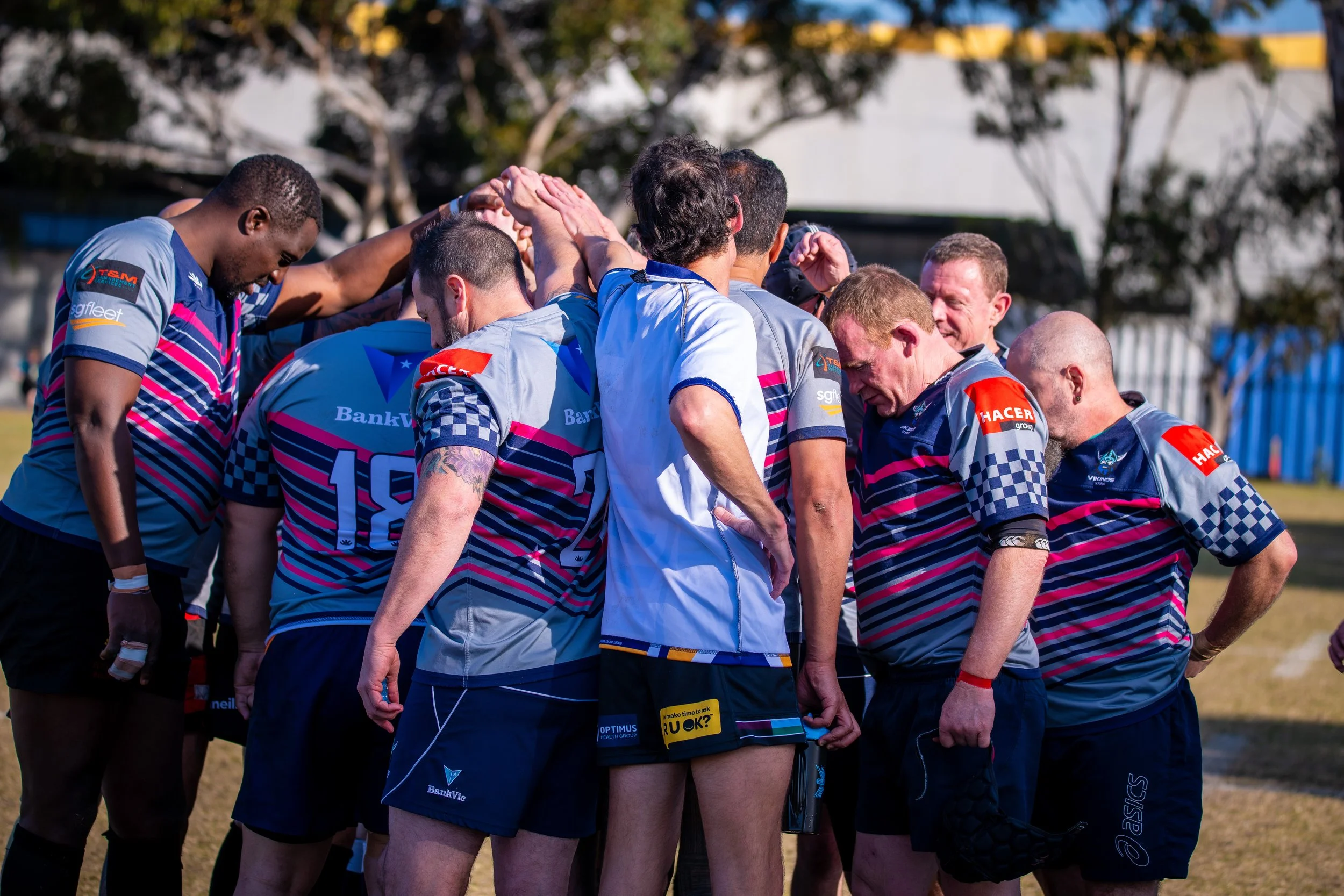 Rugby players in a team huddle on the field during daytime, wearing matching jerseys with blue and pink stripes, with a coach or team staff member.