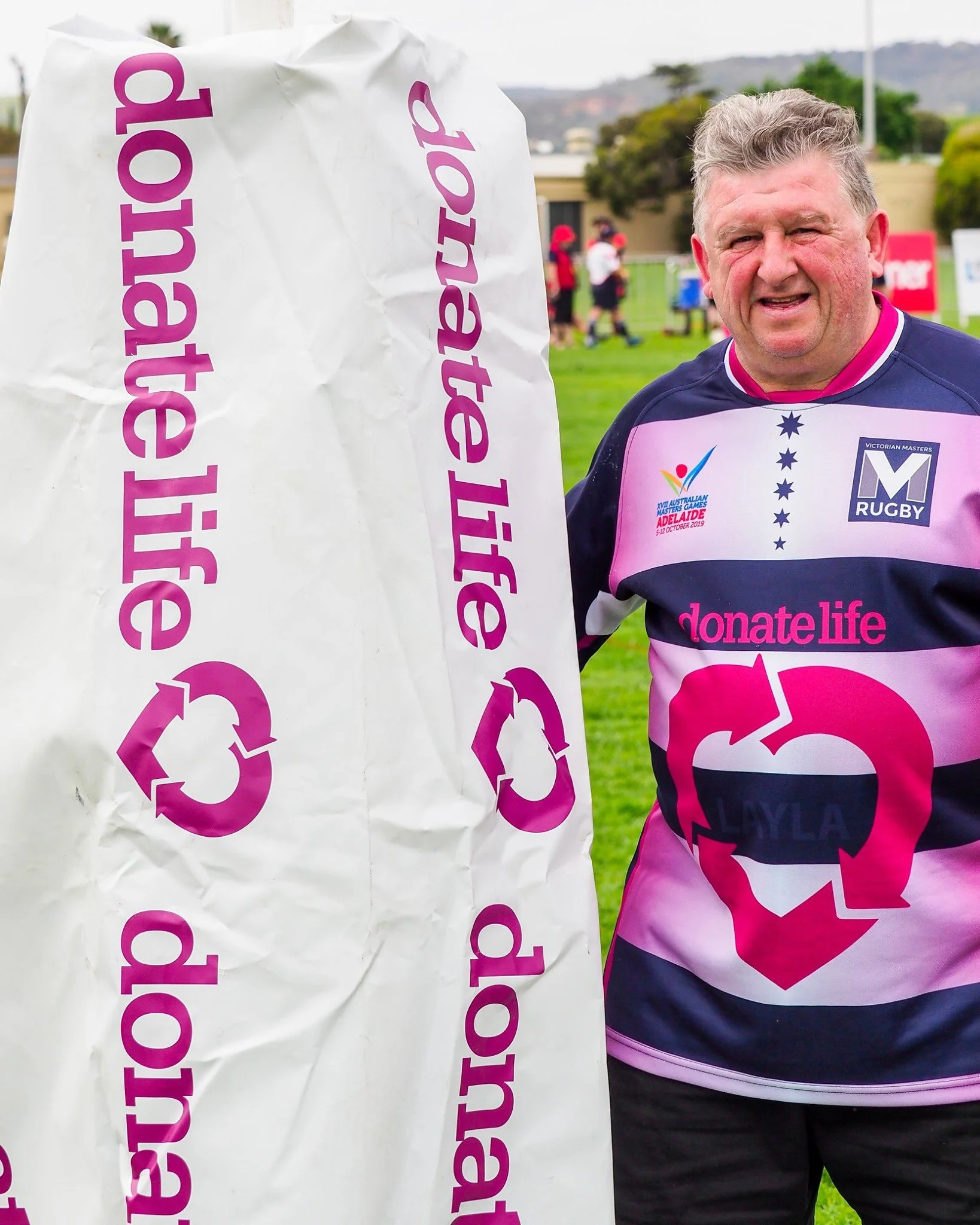 A man wearing a rugby jersey with 'donate life' campaign logo, standing outdoors on a rugby field with a large white bag marked 'donate life' and the donation symbol.