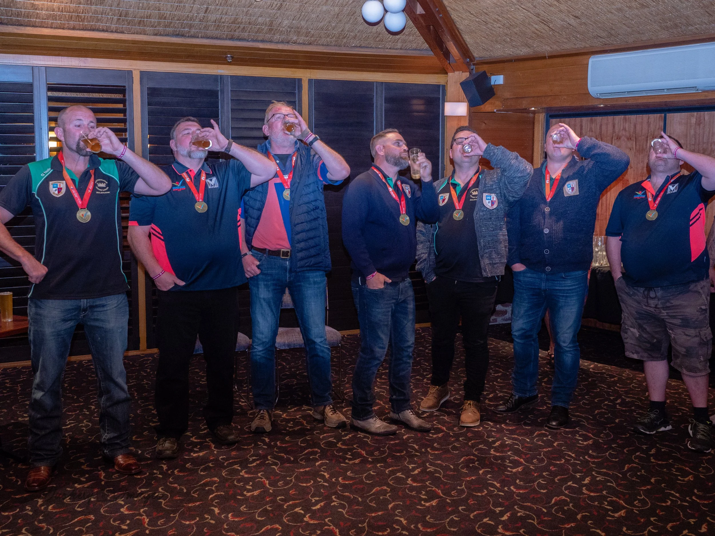 A group of eight men standing in a row drinking beer, wearing medals and team sports jerseys in an indoor setting with wooden walls and carpeted floor.