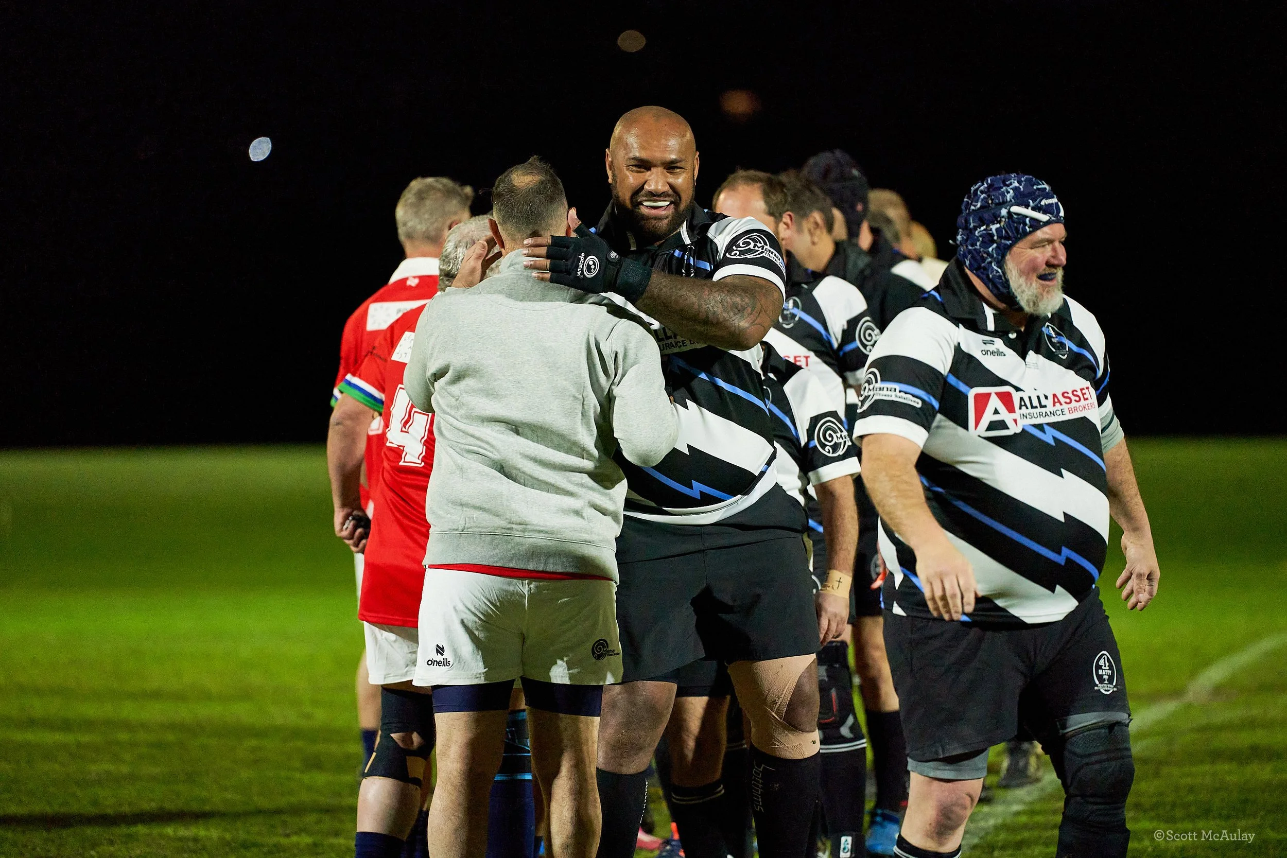 Rugby players lined up after a match, with one player in black and blue jersey hugging a person in grey, smiling. The scene is set at night on a grassy field.