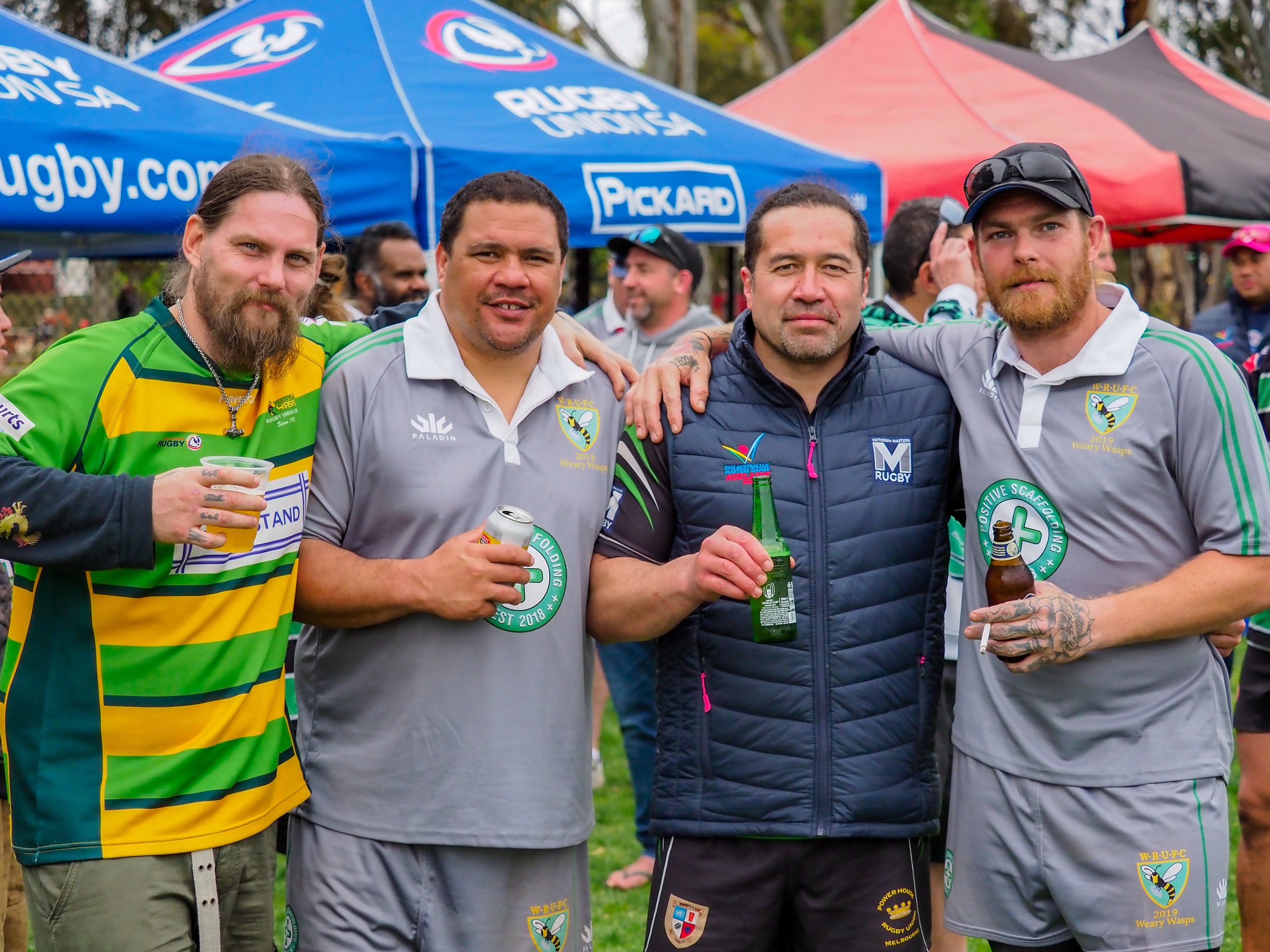 Four men standing together at a rugby event, holding drinks and smiling. They are wearing athletic and casual clothing, with some in rugby jerseys. There are tents and other people in the background.