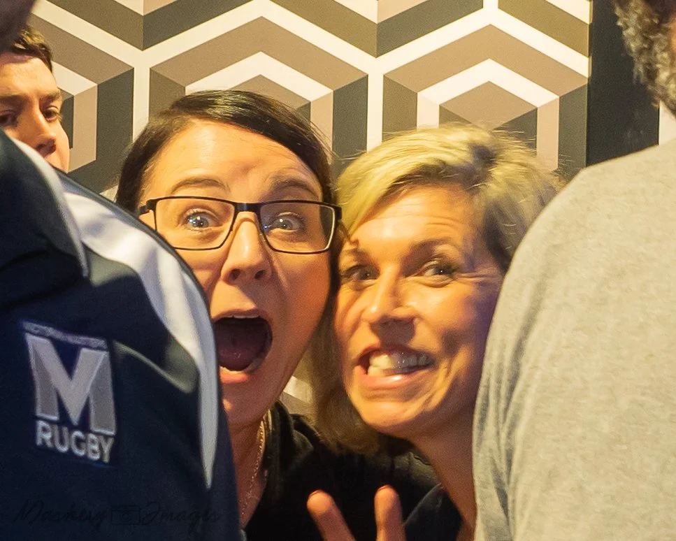 Two women smiling and making silly faces for a selfie, surrounded by a group of people indoors with a geometric patterned wall in the background.