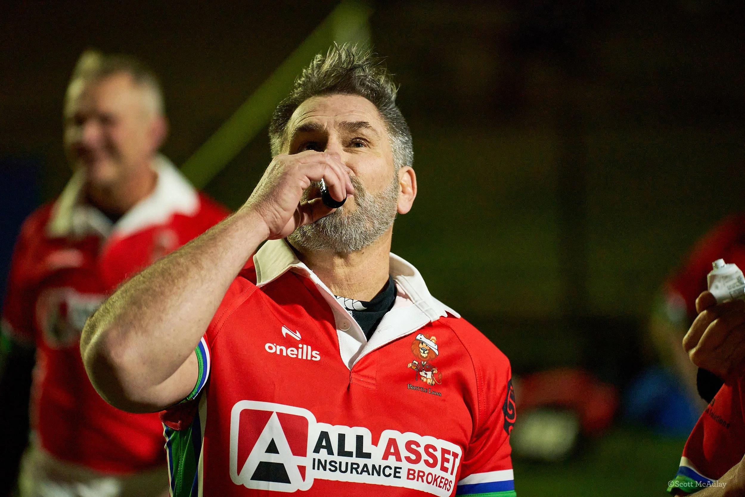 A man wearing a red rugby jersey with sponsors' logos, drinking from a small container, during a game or event at night with blurred people in the background.