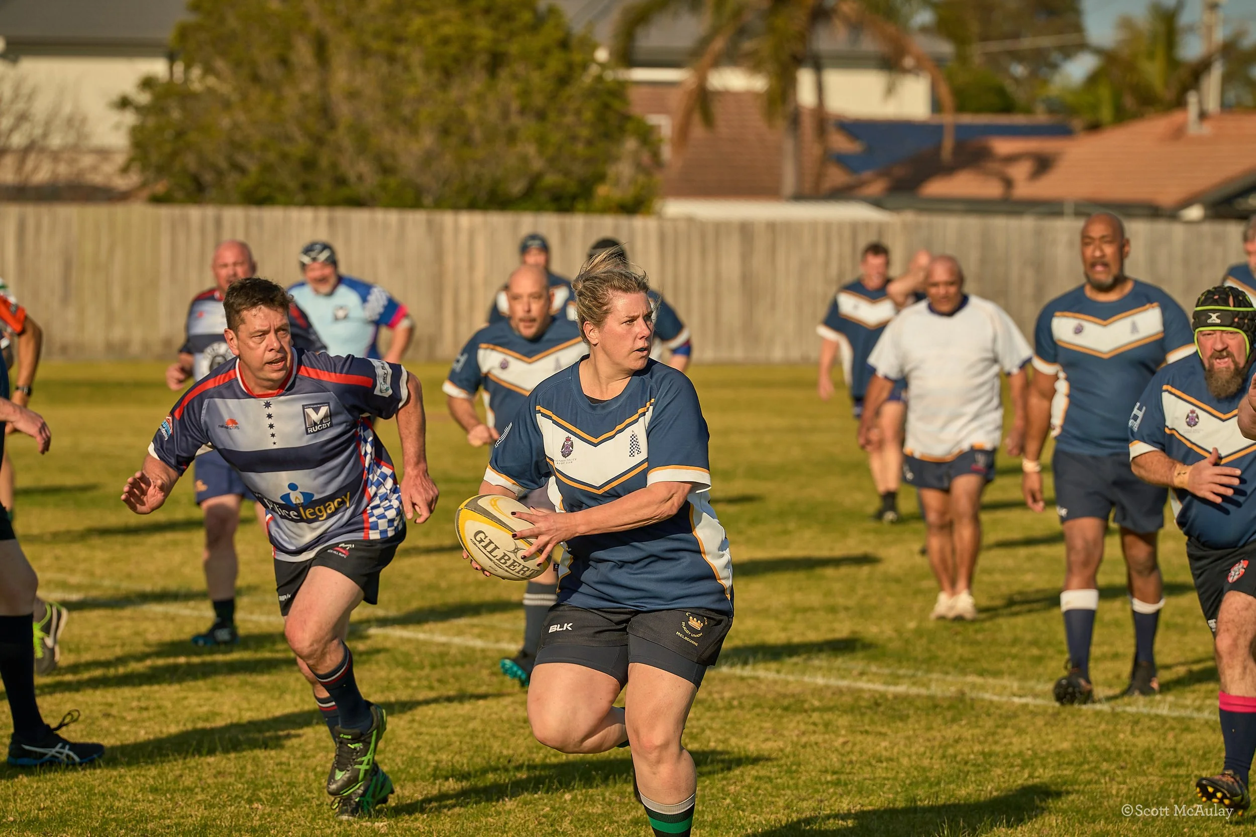 Rugby players in action on a field, with a female player holding a rugby ball, surrounded by male players. The players are wearing various jerseys and the scene occurs outdoors with houses and trees in the background.