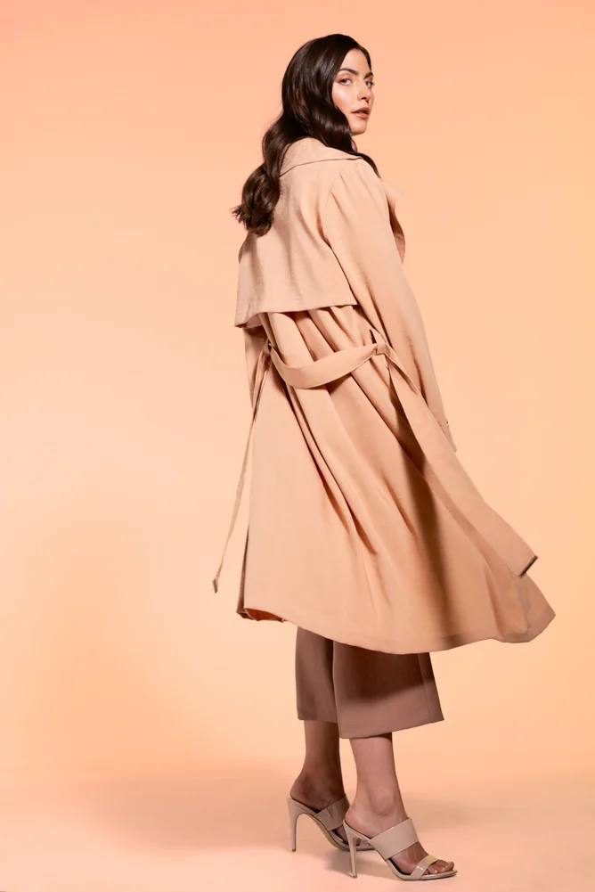 A woman with long, dark hair wearing a beige trench coat, beige pants, and high heels against a light peach background.
