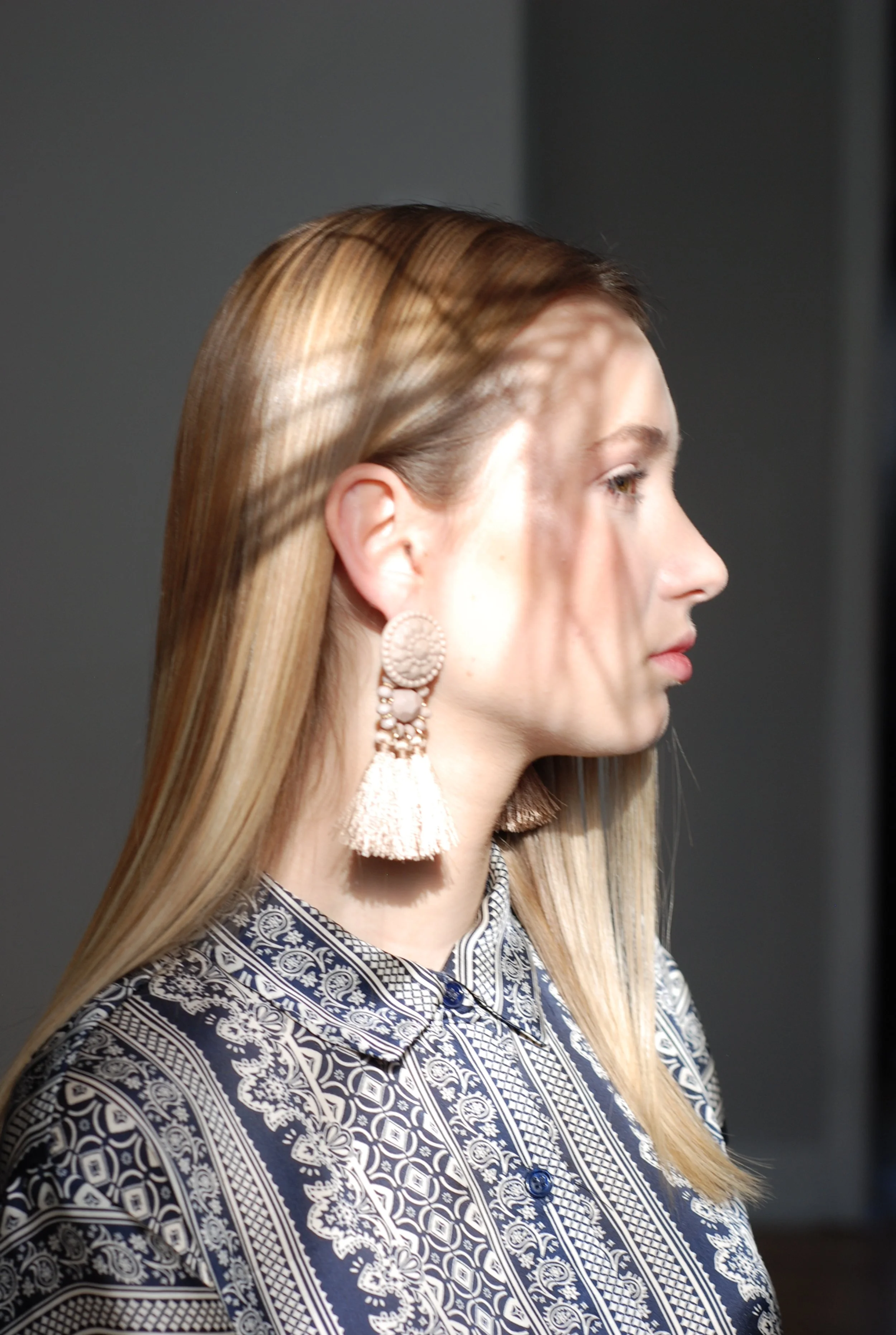 Side profile of a woman with smooth, straight blonde hair wearing a patterned shirt and large, tassel earrings.