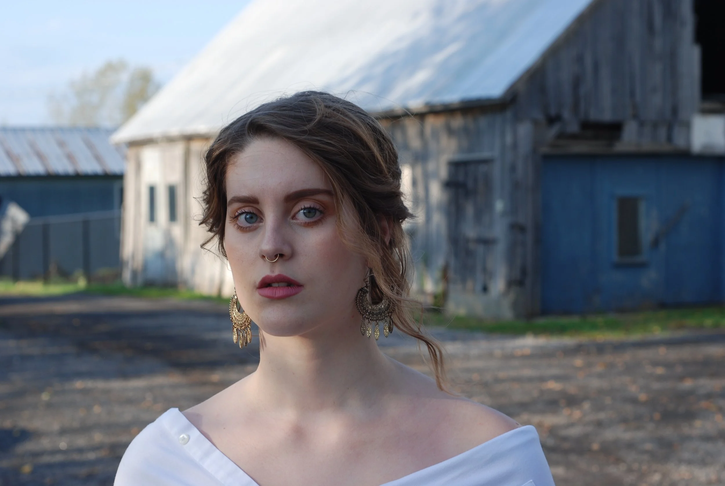 A young woman with blue eyes and short brown hair, wearing large earrings and a white off-shoulder top, standing outdoors in front of a rustic barn on a cloudy day.