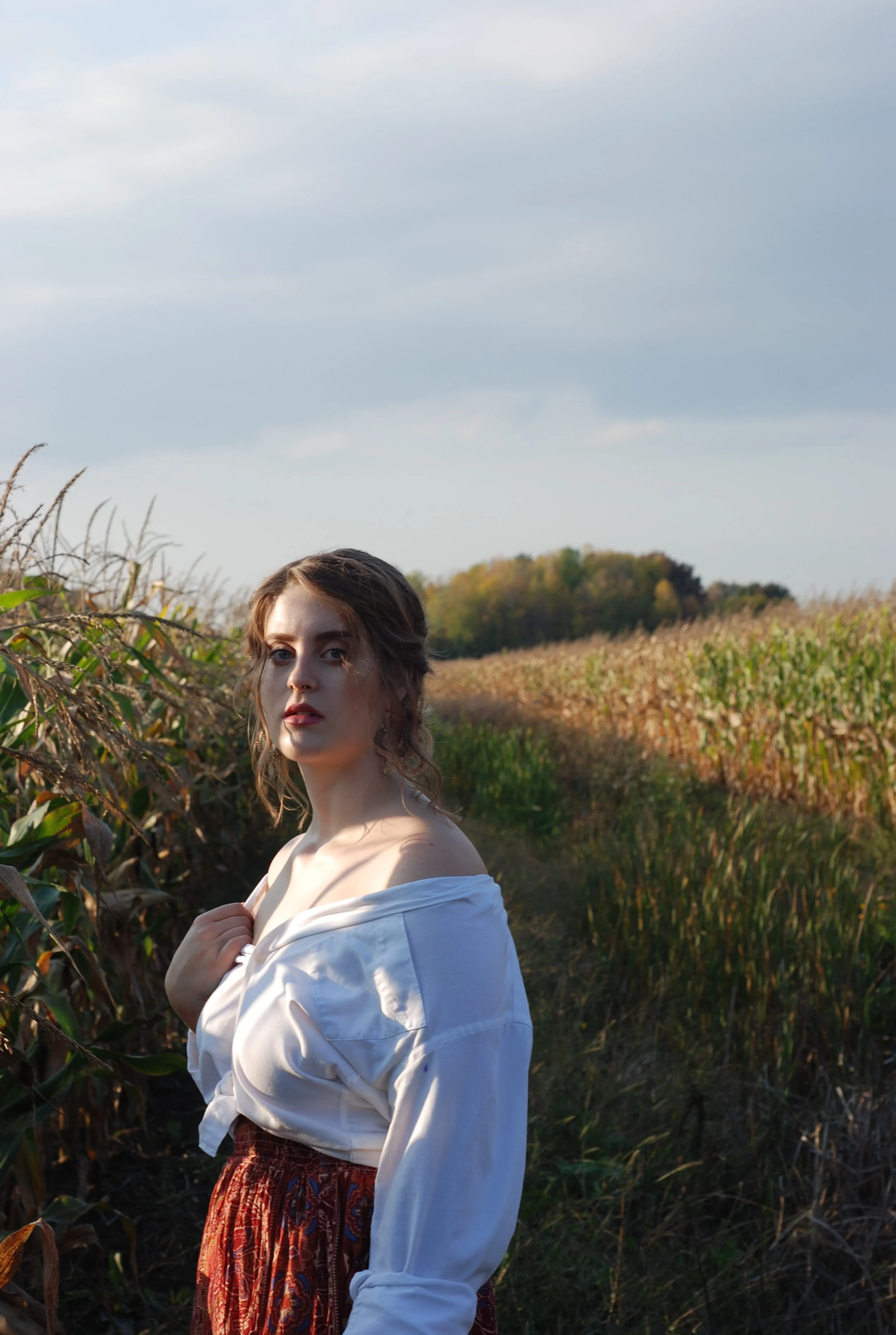 A young woman with wavy hair stands in a cornfield during late afternoon or early evening, wearing an off-the-shoulder white blouse and a patterned skirt, looking at the camera with a serious expression.