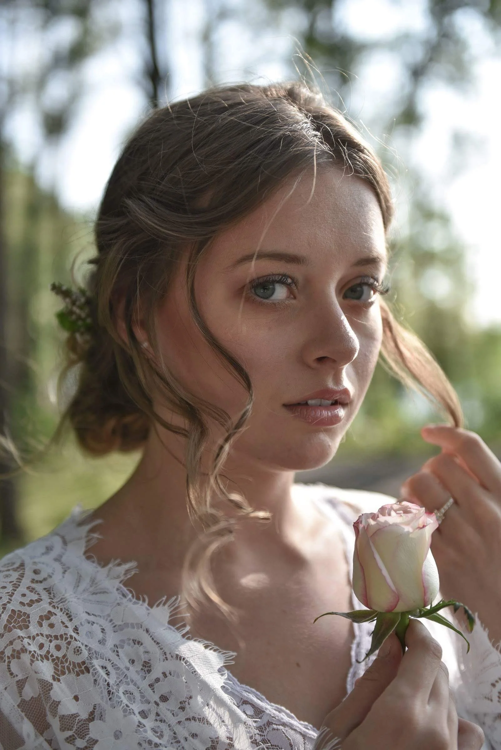 A young woman with wavy brown hair and blue eyes holding a pink-and-white rose, wearing a white lace top, outdoors with blurred trees and greenery in the background.