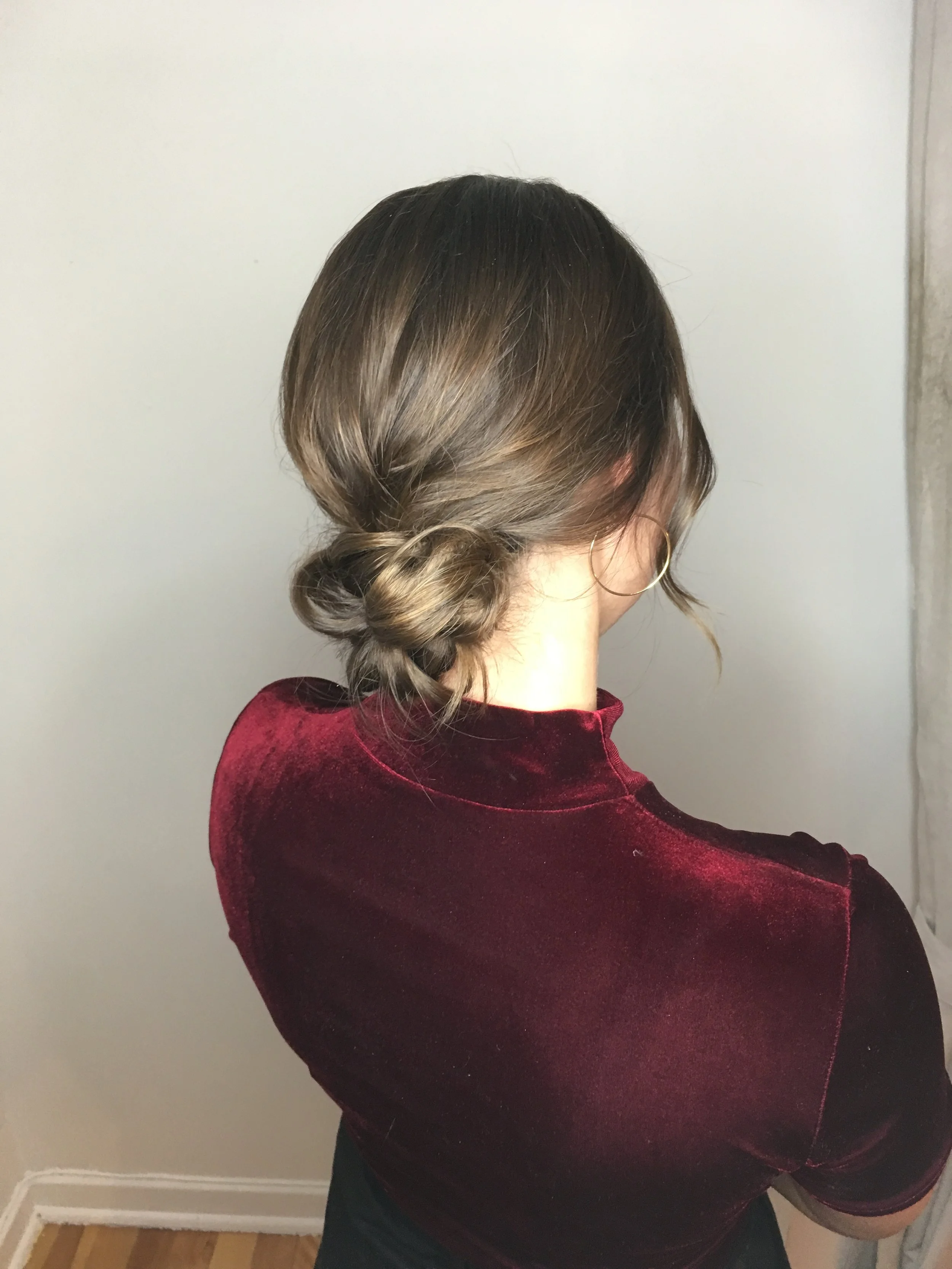Woman with brown hair styled in a low twisted bun, wearing a burgundy velvet top and hoop earrings, standing indoors with a plain white wall in the background.