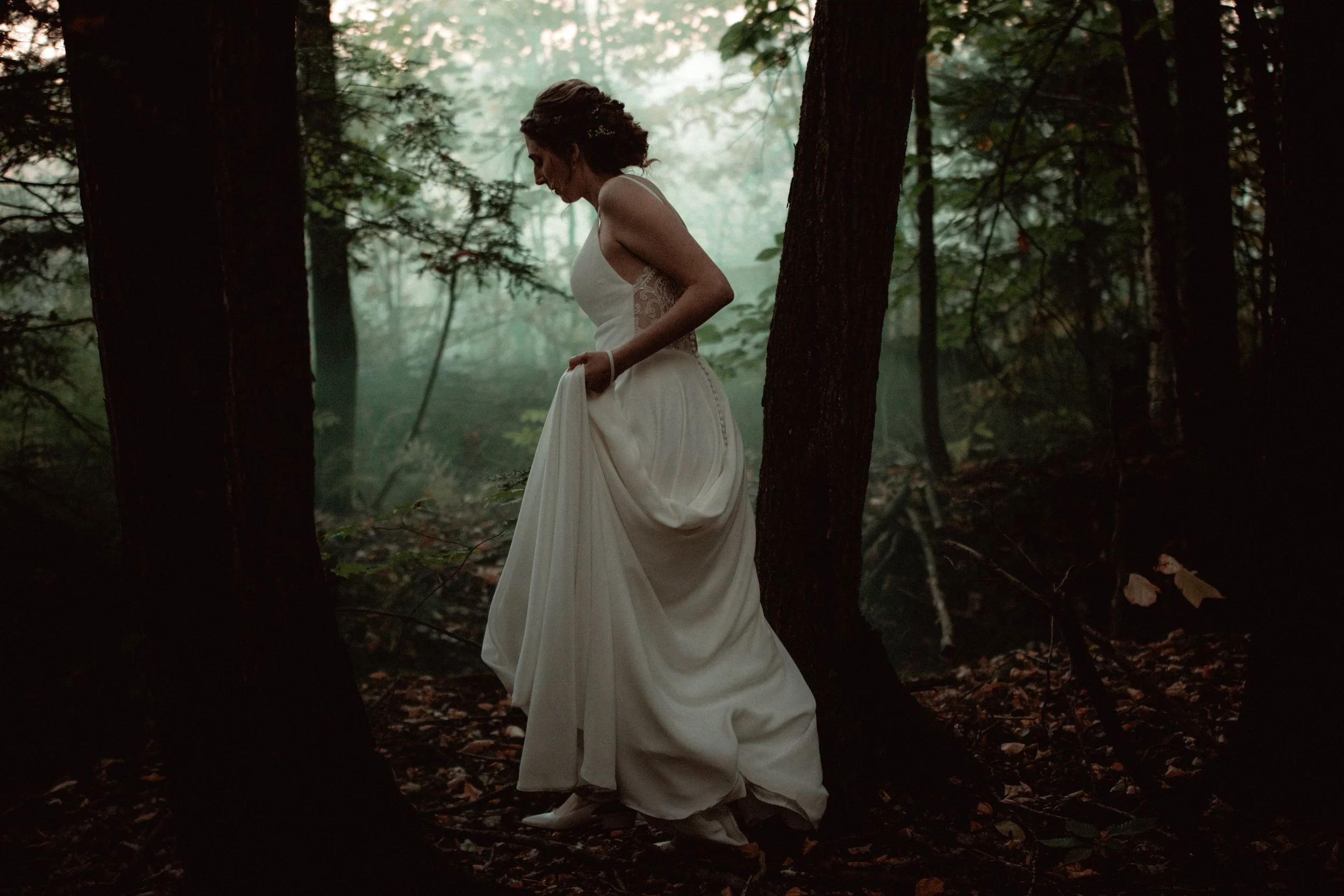 A woman in a white wedding dress standing in a dark, foggy forest, looking down and holding part of her dress.