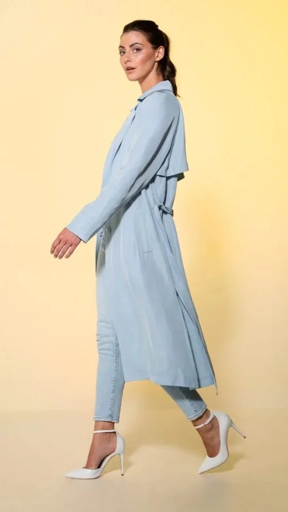 A woman with dark hair tied back, wearing a light blue trench coat, light blue jeans, and white high heels, walking against a yellow background.