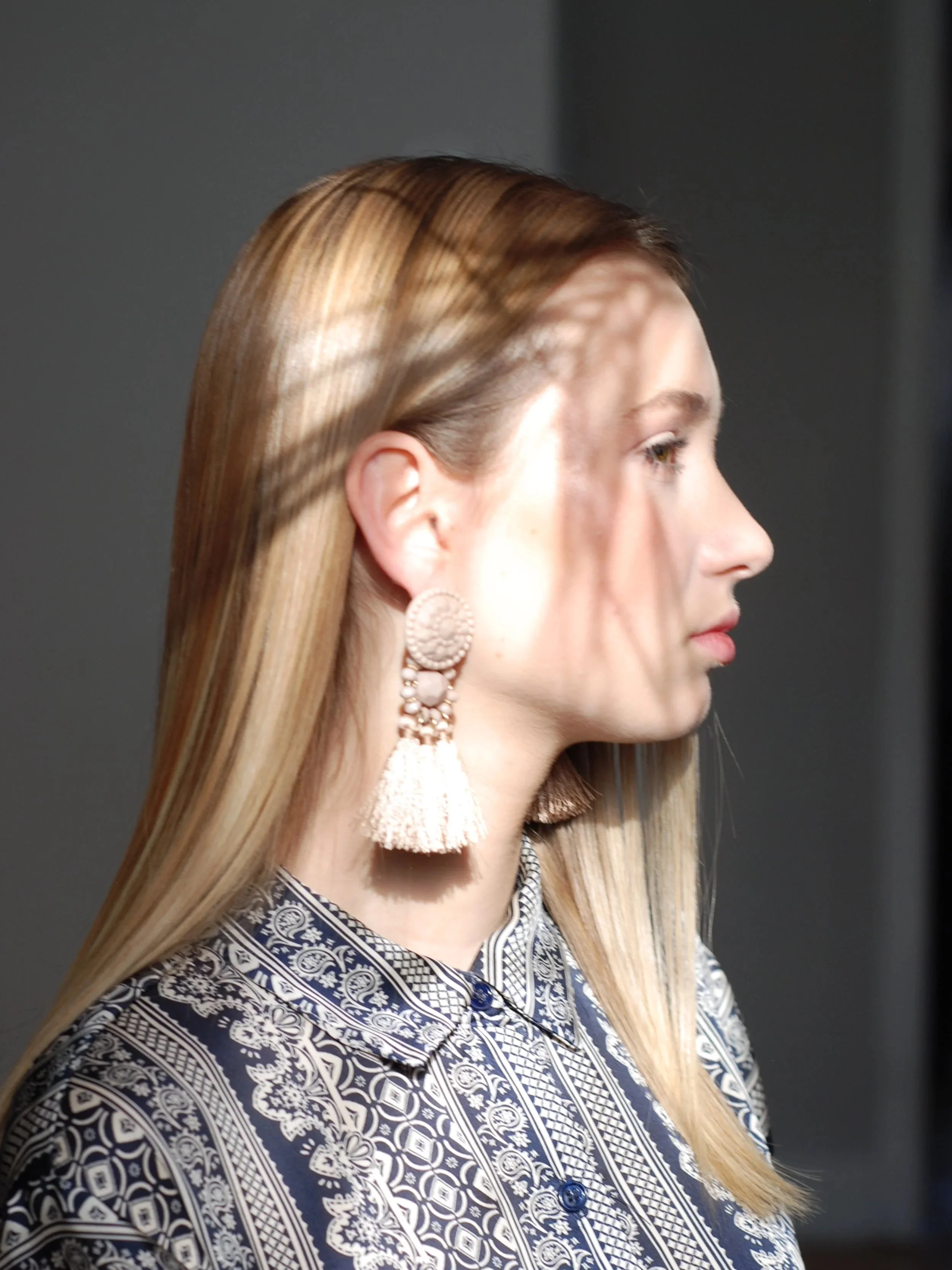 Side profile of a woman with smooth, straight blonde hair wearing a patterned shirt and large, tassel earrings.