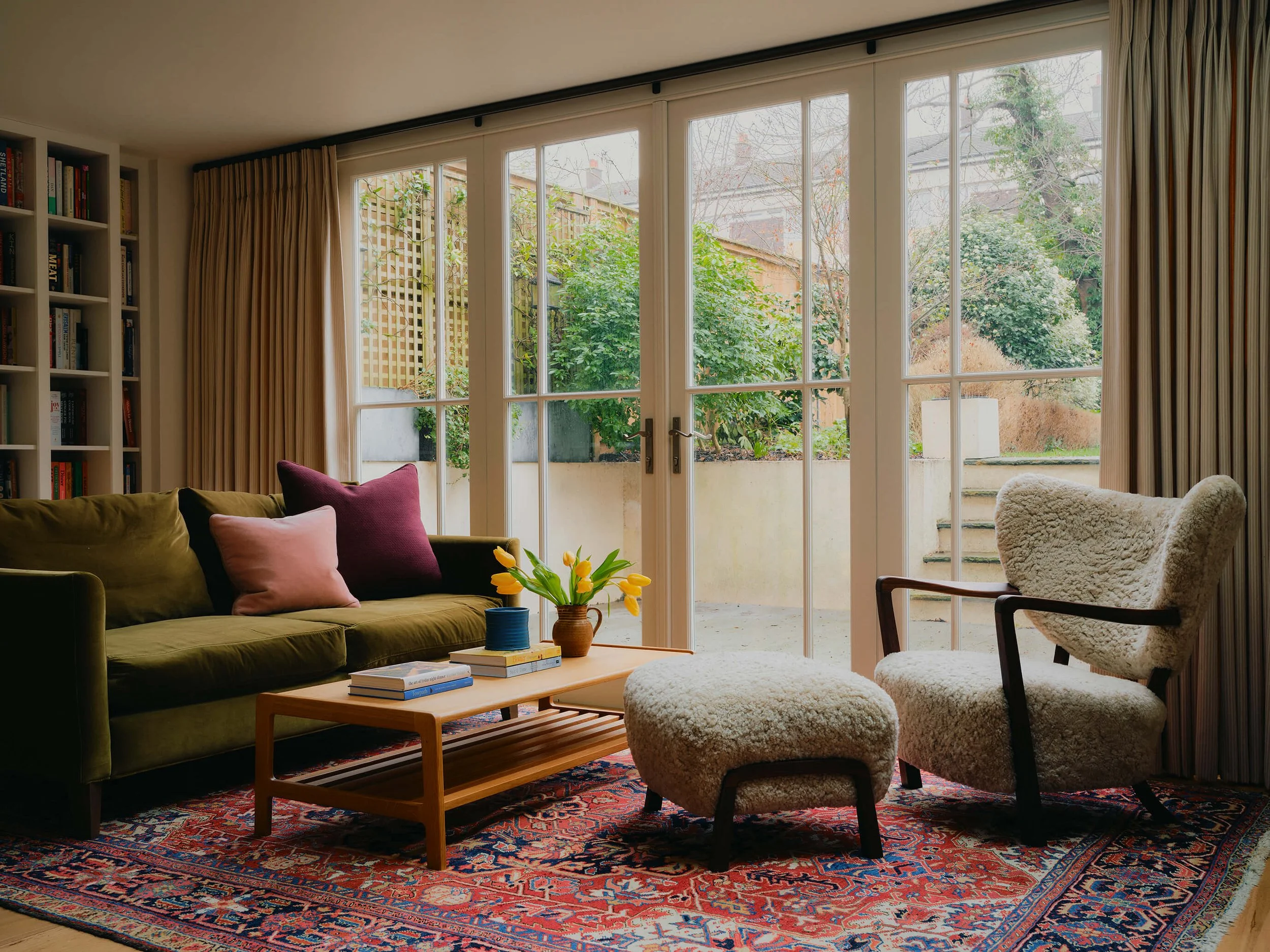Living space with doors out to the garden. Green velvet sofa, wooden coffee table, ornate rug, and filled bookshelves.