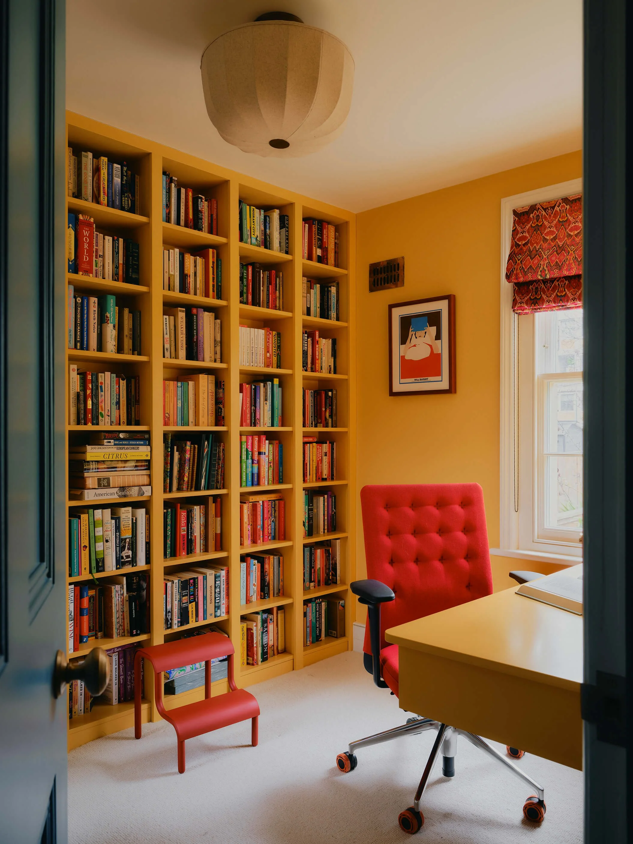 Bright yellow study, one wall is a fully filled book shelf, the room has a yellow desk and bright red office chair.