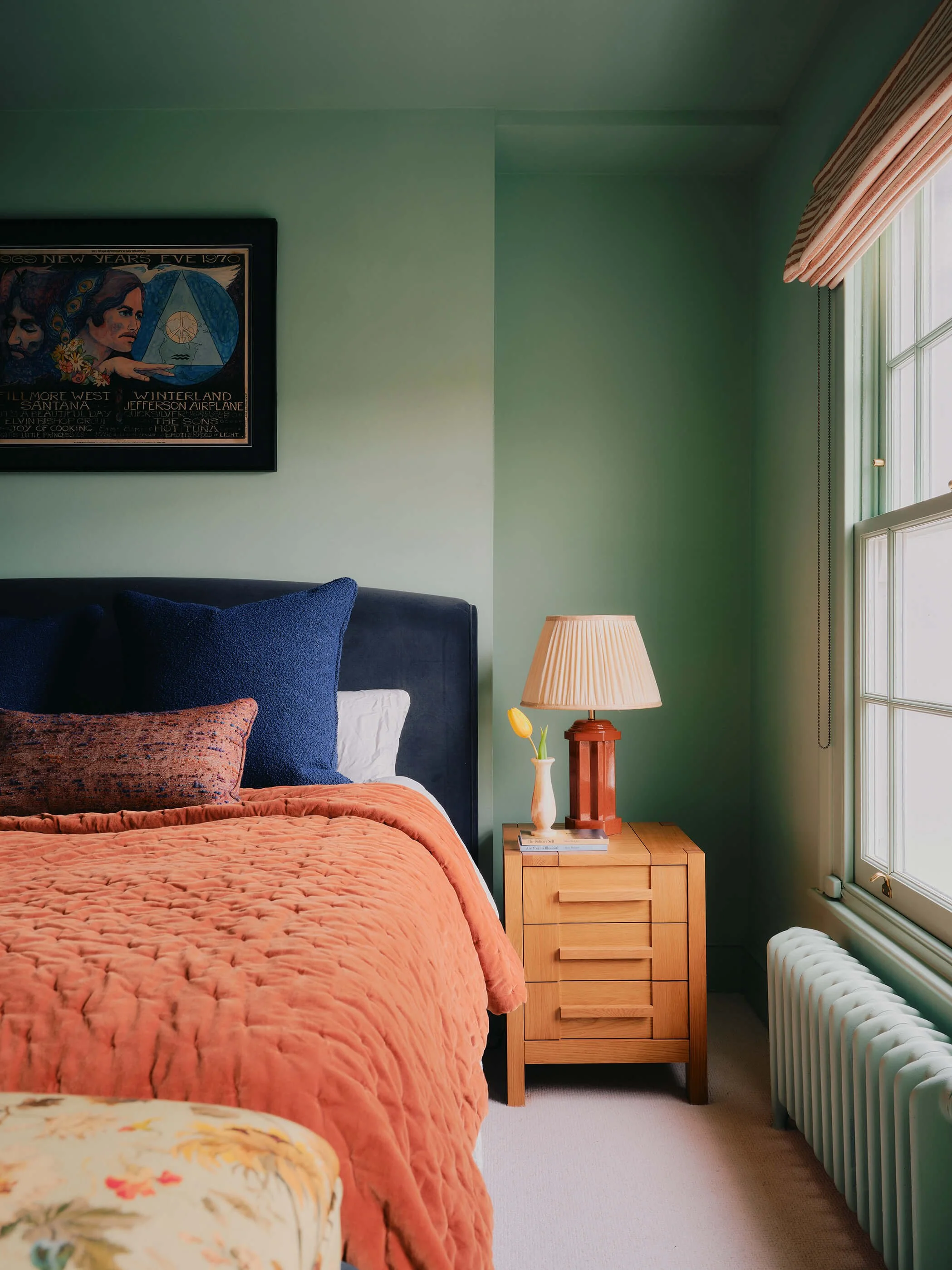 Mint green painted bedroom with orange and blue bed, wood sidetable with lamp and single tulip.