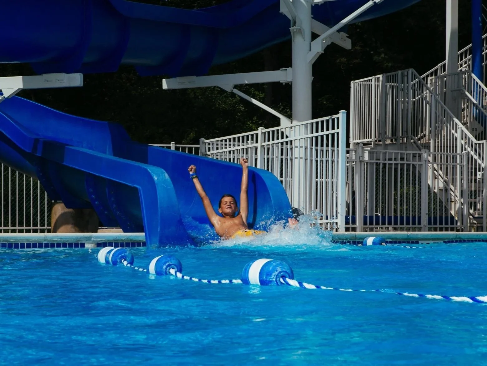 Child joyfully sliding down a blue water slide into a swimming pool with water splash, surrounded by white safety fencing at a water park.