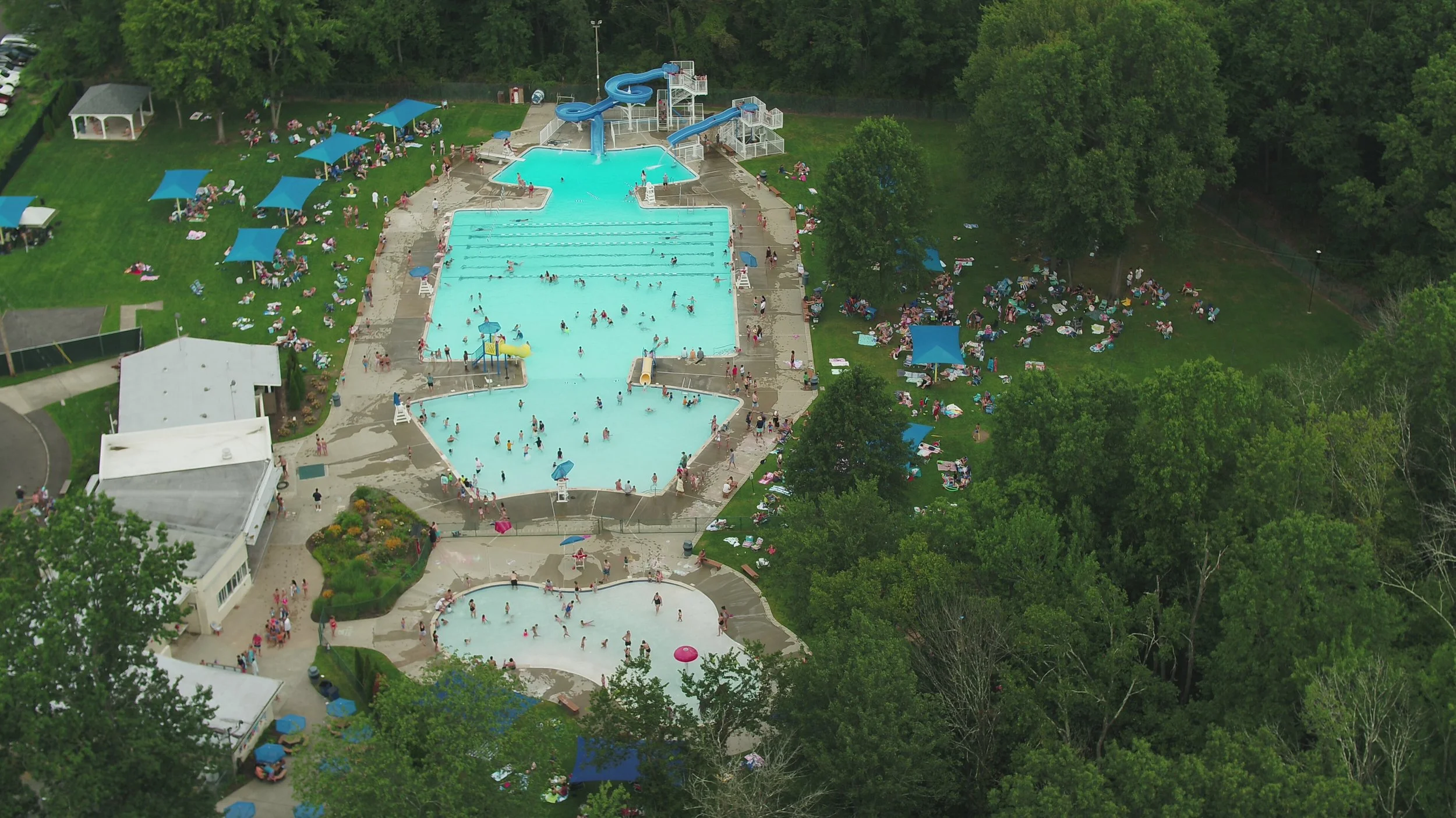 An aerial view of a crowded public swimming pool area surrounded by green trees, with several pools, water slides, and many people swimming and relaxing on the grass with umbrellas.