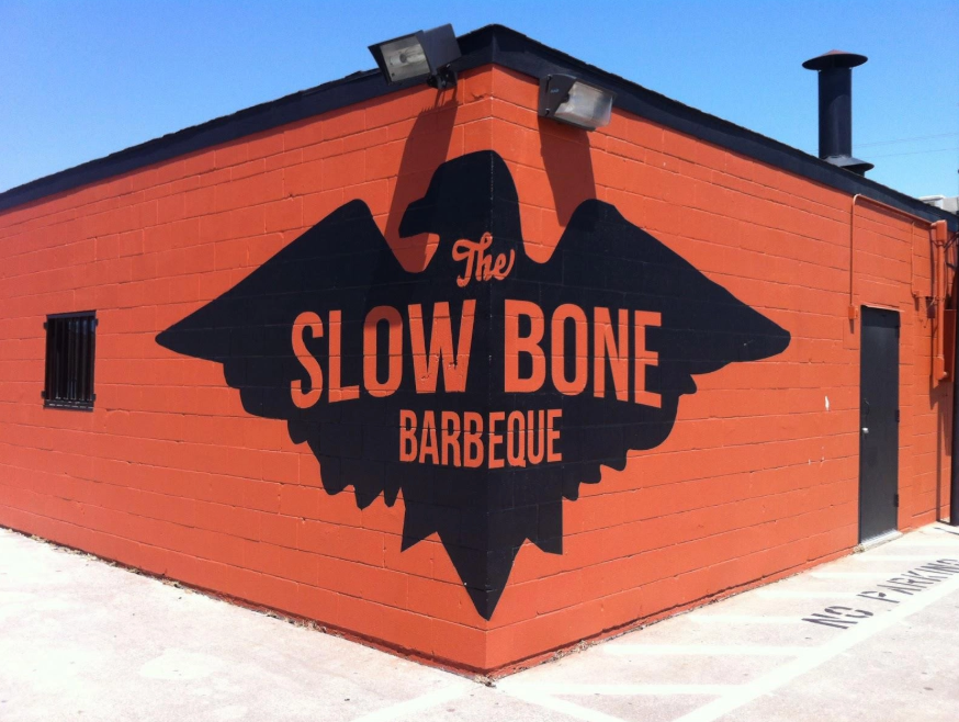 A red brick building with a black logo on the side that reads "The Slow Bone Barbeque" in bold orange letters, with a silhouette of a pig's head above the text.