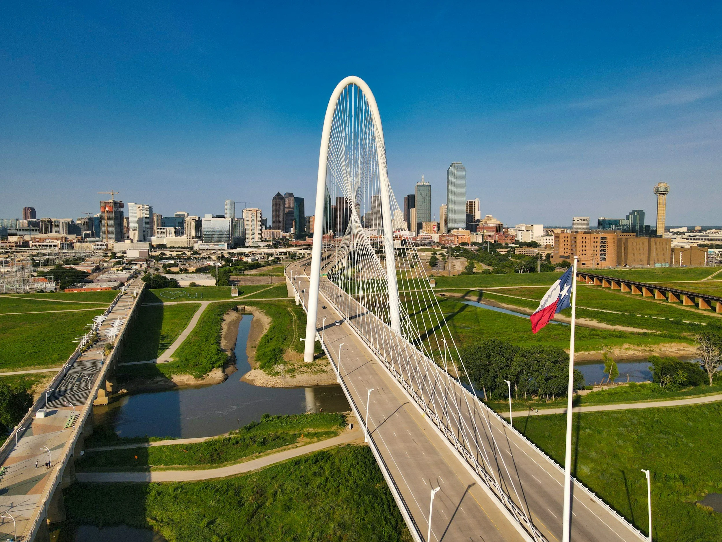 An aerial view of a city skyline featuring a large modern arch bridge over a river, with a Texas flag flying in the foreground, and tall buildings in the background under a clear blue sky.