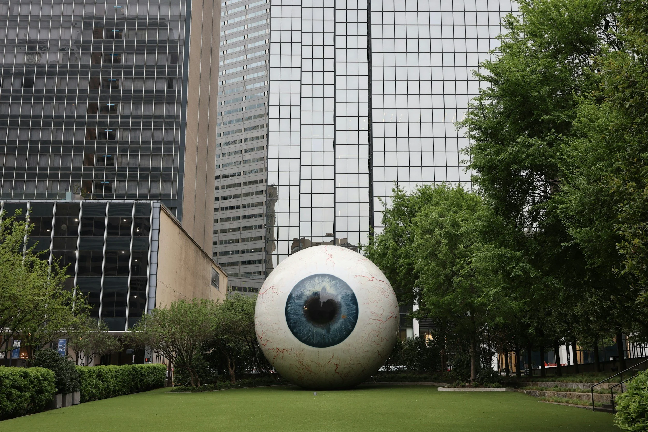 A large sculpture of a human eyeball situated on a grassy area surrounded by bushes and trees, with tall office buildings and a glass skyscraper in the background.