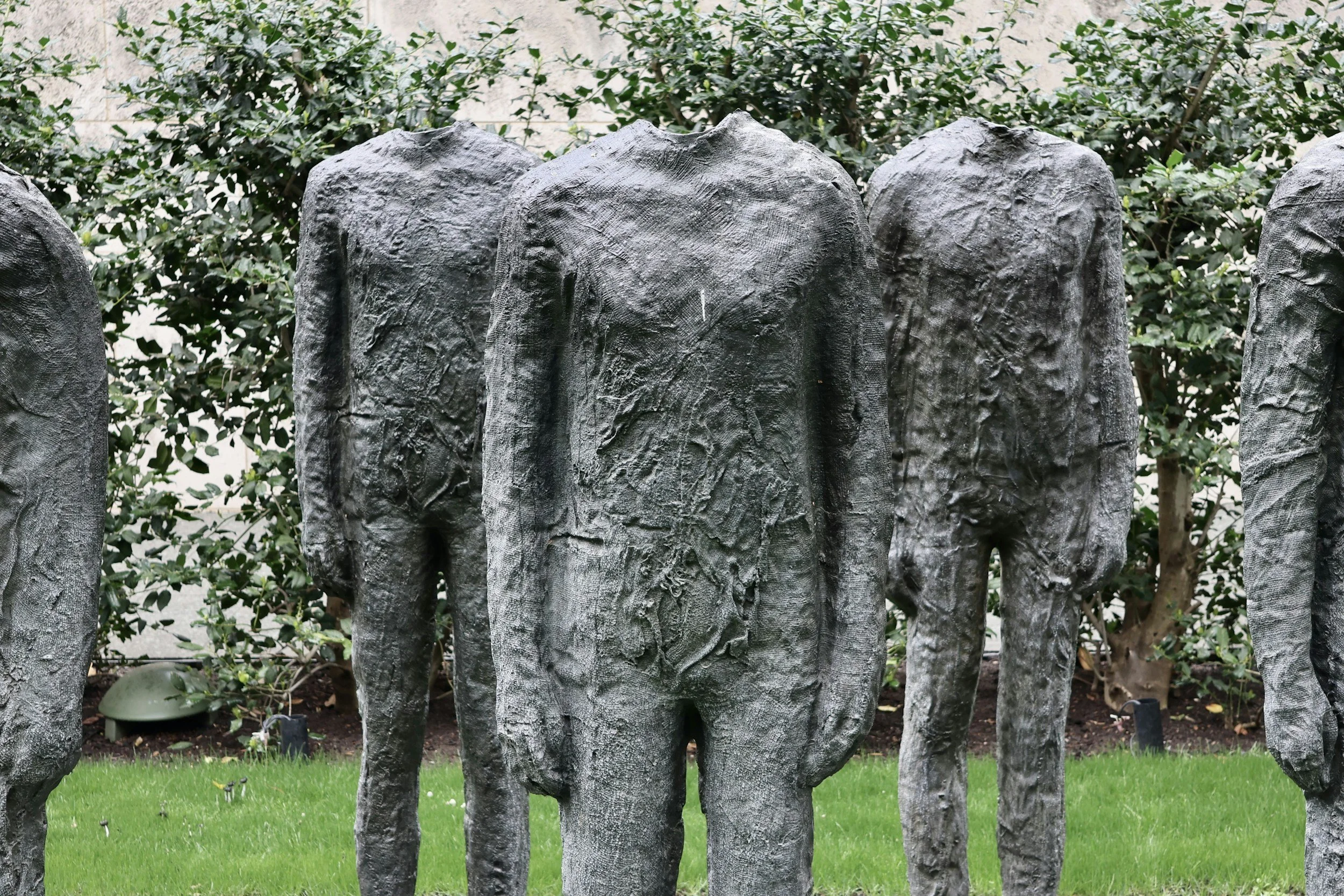 Several bronze statues of faceless, headless figures standing outdoors on grass, with bushes in the background.