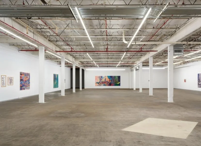 An art gallery with white walls displaying colorful paintings, a high ceiling with exposed pipes and lighting, and a large open floor space.