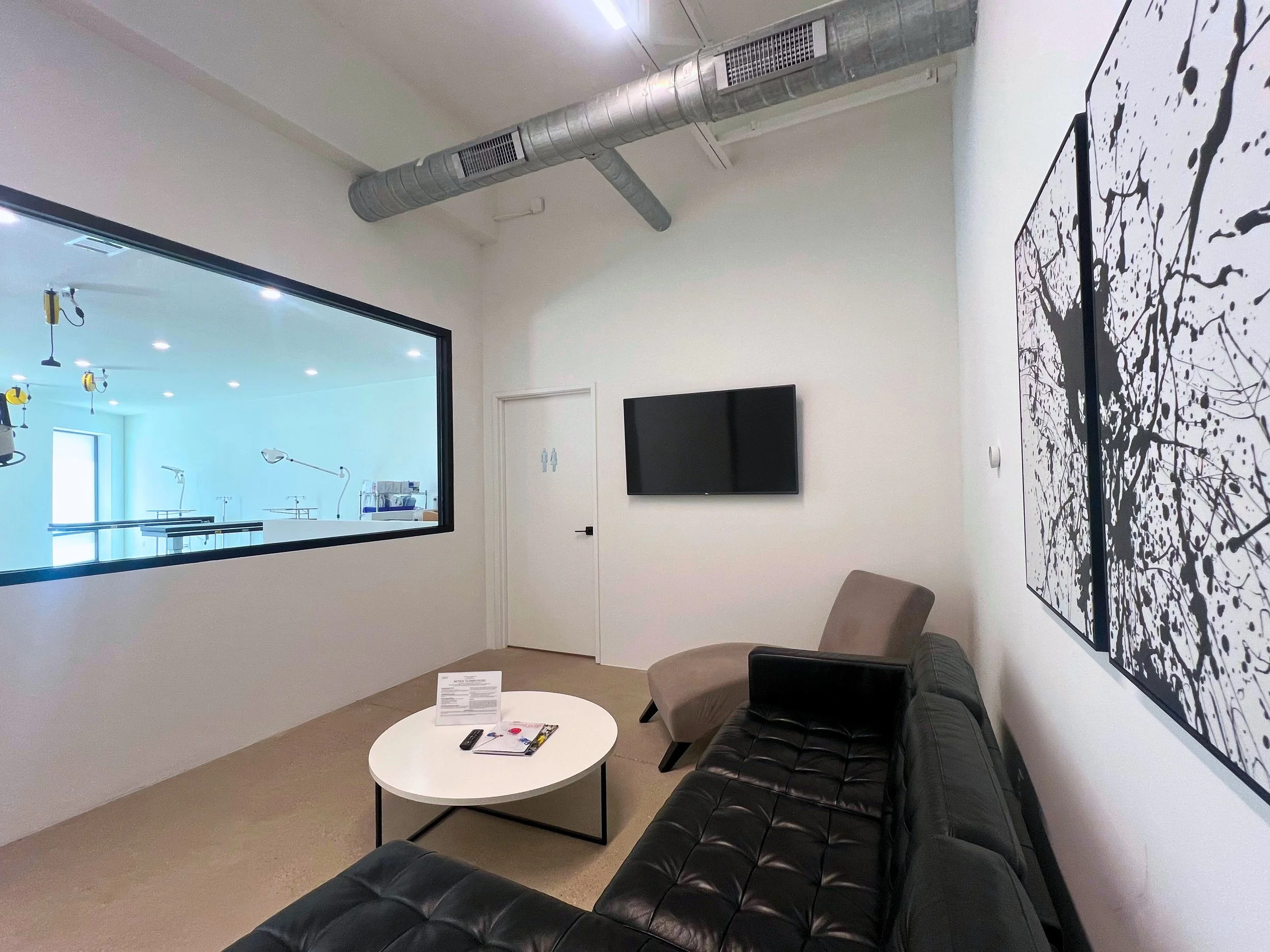 A modern medical or hospital waiting room with a black leather sofa, a beige armchair, a white round coffee table with papers and remote on it, a wall-mounted TV, a large painting with black splatter art, a window to an operating room or hospital procedure area, and an exit door with restrooms symbol.