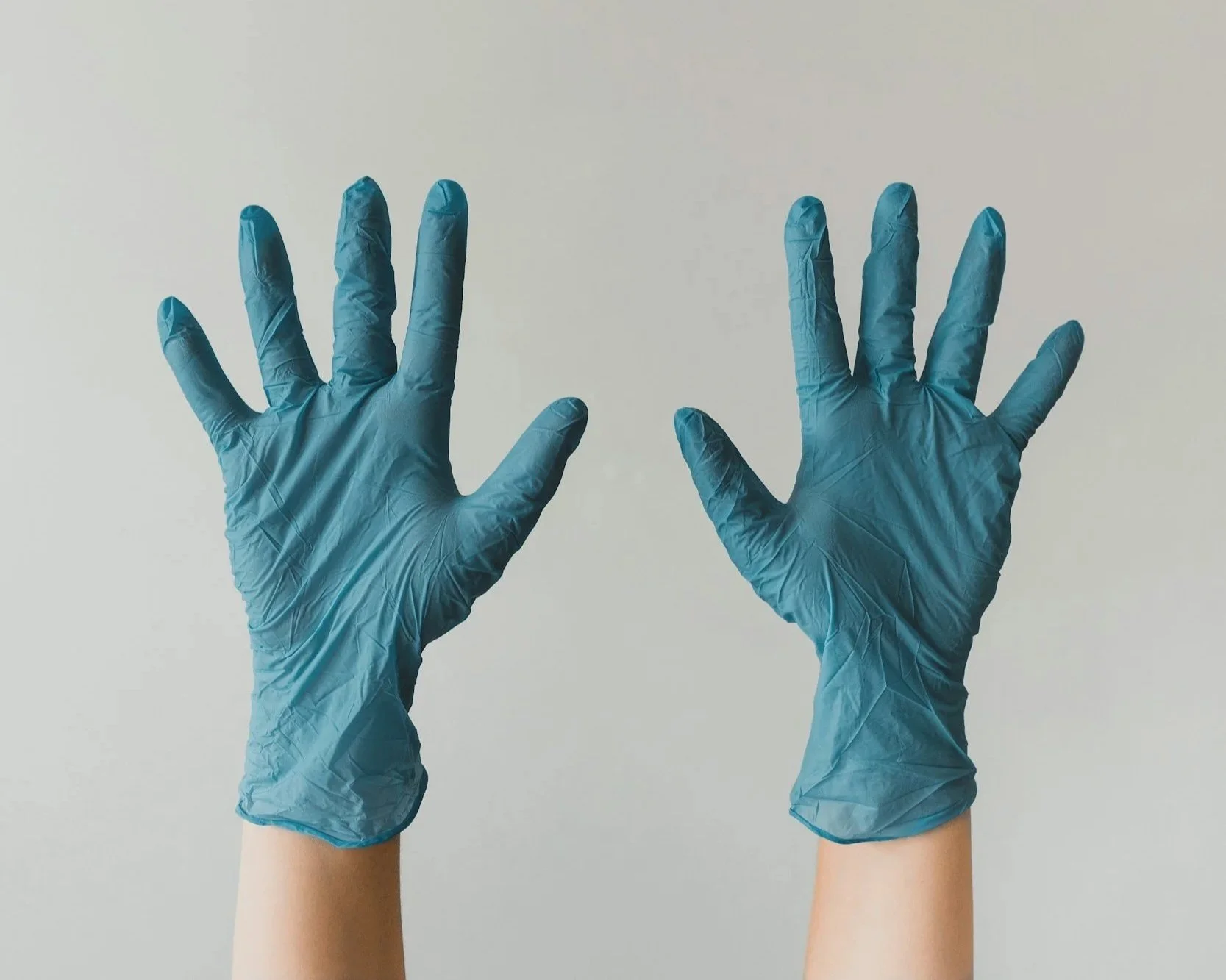 Two hands wearing blue medical gloves raised against a plain background.