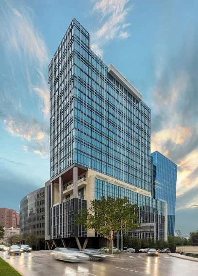 Modern glass high-rise building with several floors, situated on a city street with cars and trees, under a partly cloudy sky during sunset.