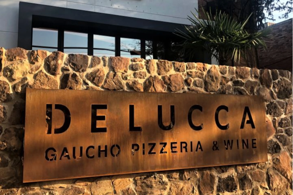 A metal sign with the text 'DELUCCA GAUCHO PIZZERIA & WINE' mounted on a stone wall, with a modern building and some greenery visible in the background.