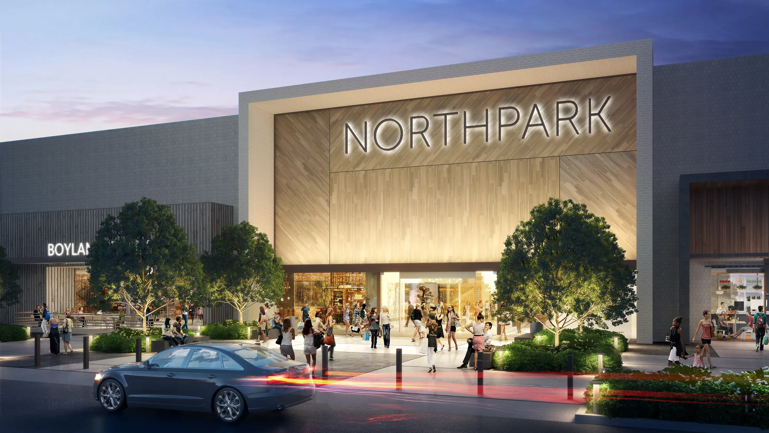 Exterior view of Northpark shopping mall during evening with the sign illuminated, people walking and sitting outside, trees, and a moving car on the street.