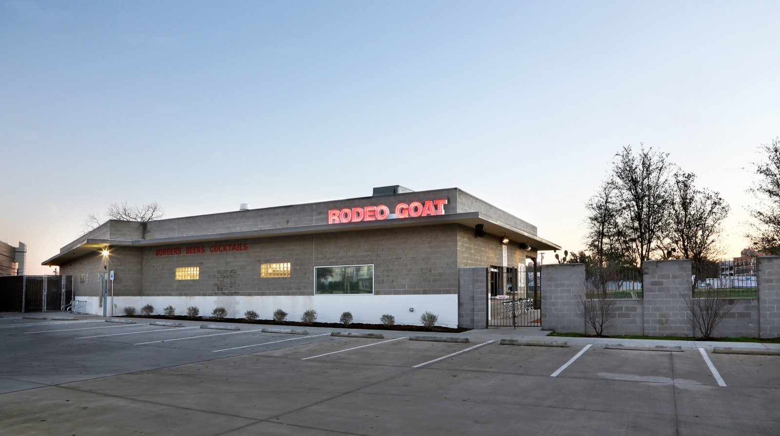 A single-story restaurant with a neon sign that reads 'RODEO GOAT' on the roof, located in a parking lot during evening with mostly clear sky, some small trees and a wall surrounding the outdoor area.