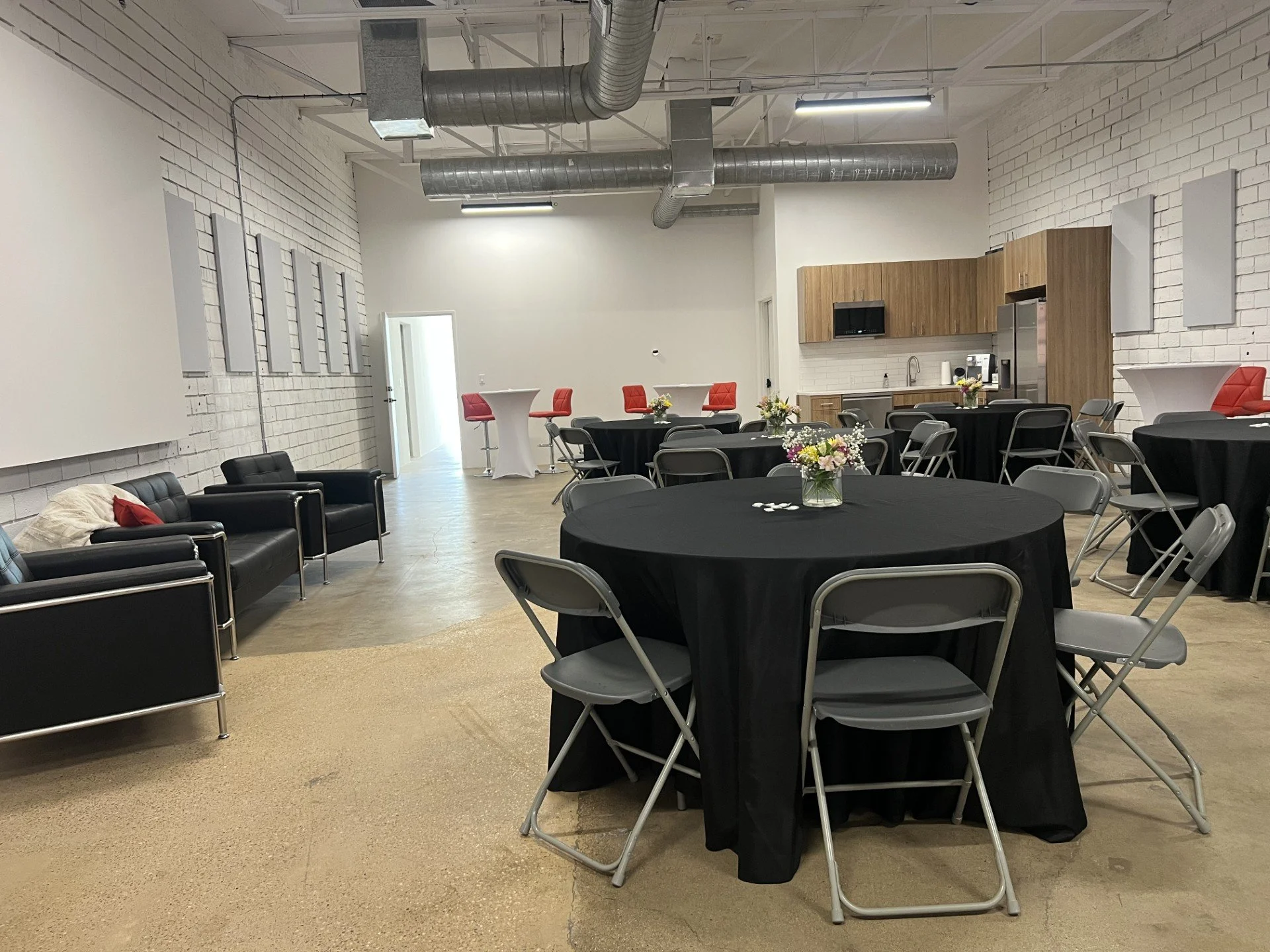 An event space with round tables covered in black tablecloths, each decorated with a small floral centerpiece. There are folding chairs around the tables, a seating area with black couches and chairs on the left, a kitchen area with wooden cabinets, a refrigerator, microwave, and coffee maker on the right, and some high-top tables with red chairs near the back wall. Exposed ductwork is visible on the ceiling, and the space has white brick walls and concrete flooring.