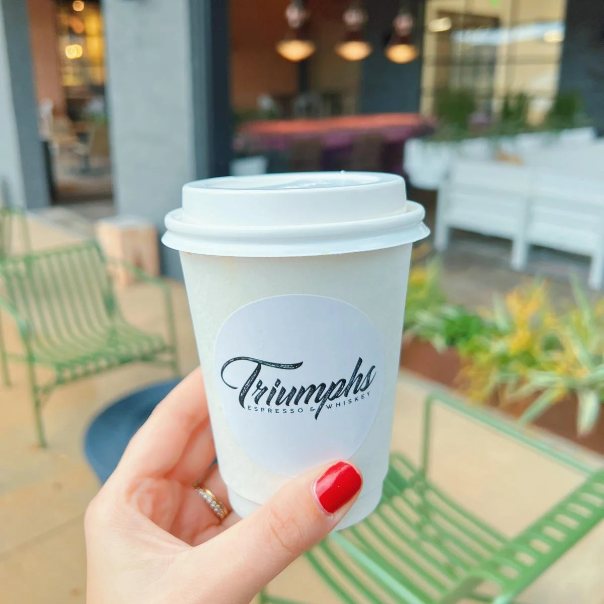 A hand holding a white disposable coffee cup with a white lid, featuring the Triumph logo, outside a cafe with green chairs and potted plants.