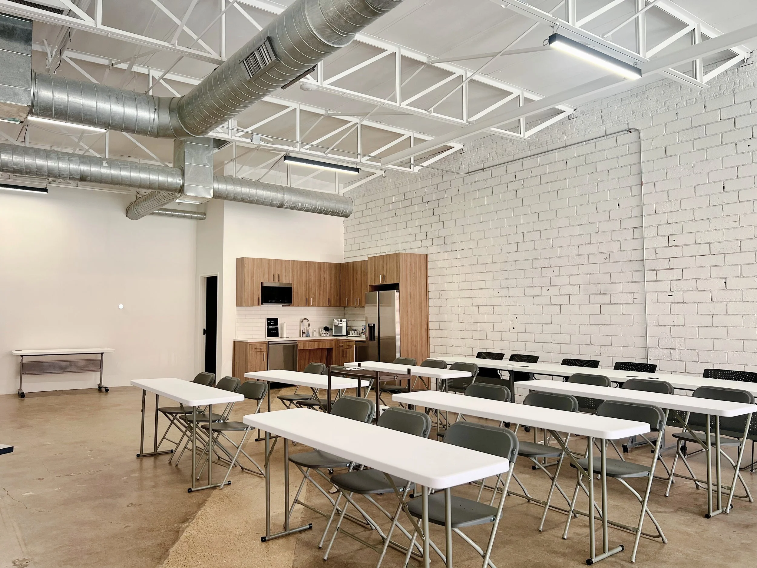 A large, empty room with white brick walls, exposed ductwork, and white metal ceiling framework. There are several white tables and gray chairs arranged in rows. In the corner, there is a kitchenette with wooden cabinets, a microwave, coffee maker, and refrigerator.