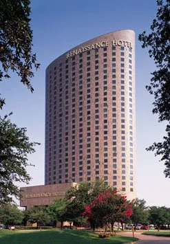 The Renaissance Hotel, a tall cylindrical building with many windows, situated in a park-like area with trees and pink flowering bushes in the foreground.