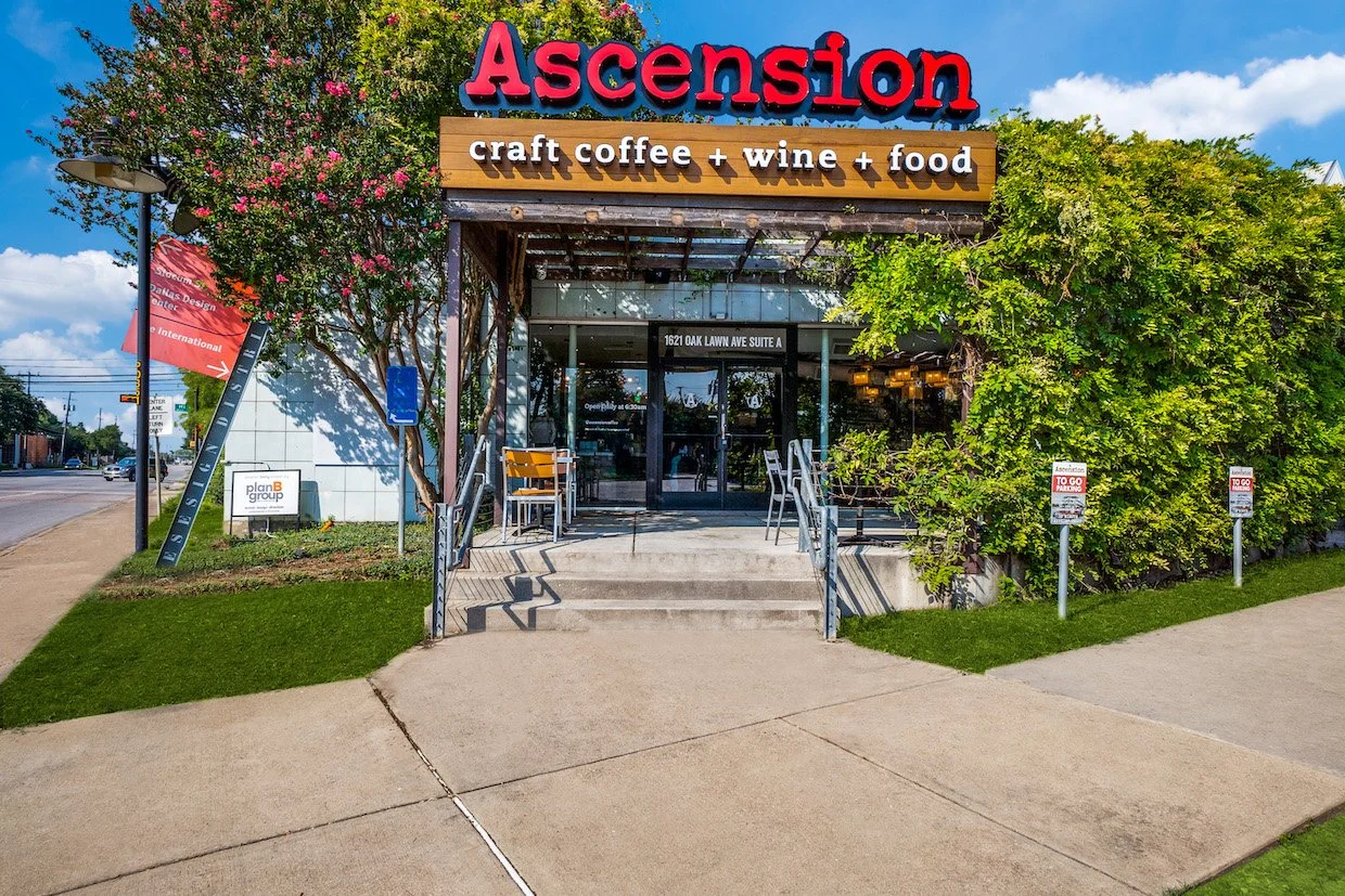 The entrance of a restaurant called Ascension with a sign that says craft coffee, wine, and food, surrounded by trees and bushes, with a sidewalk and a street to the left.