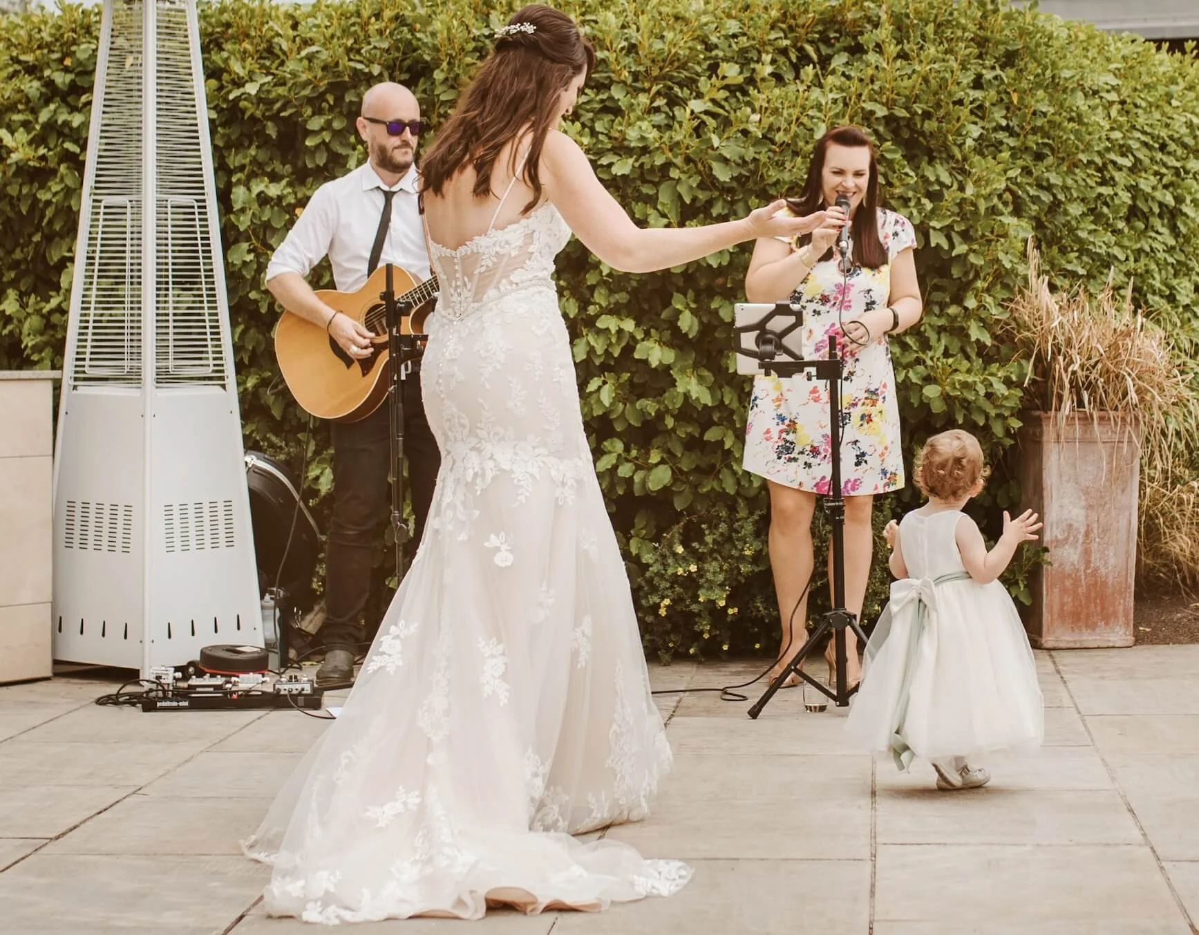 A bride in a white wedding gown dances with a young girl in a white dress, while Soulstar Music's acoustic duo plays guitar and sings at an outdoor wedding reception.
