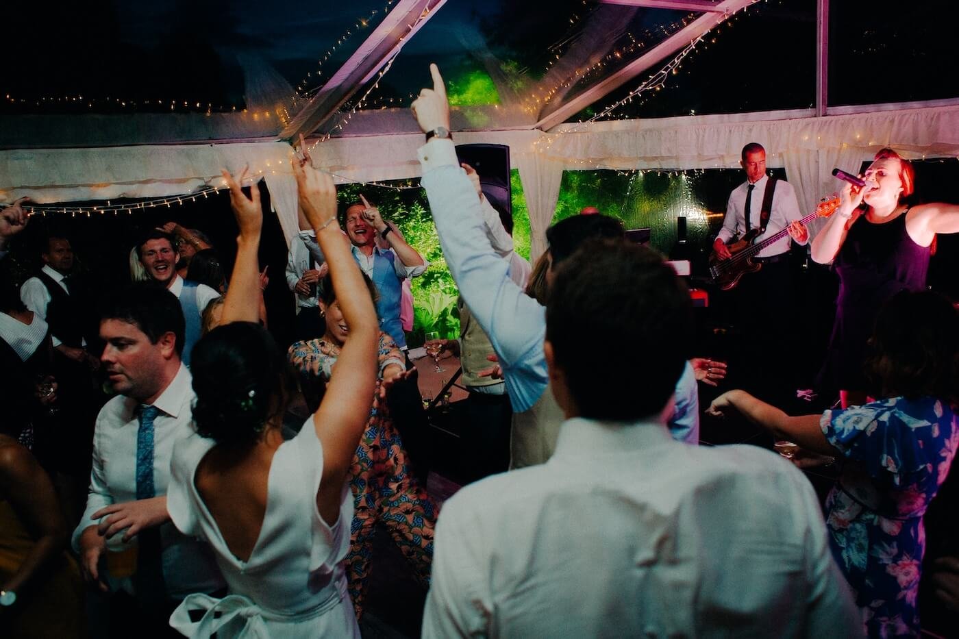 People dancing and singing at a lively wedding party with Soulstar Music live band performing on stage under a glass tent decorated with string lights.