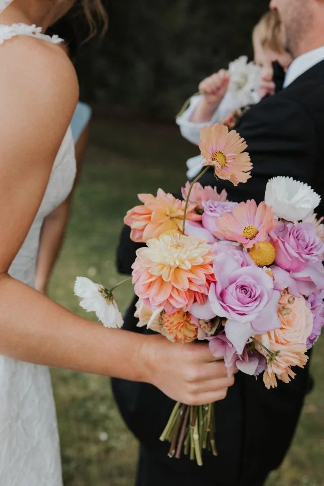 Close-up of a woman in a white dress holding a colorful bouquet of pink, orange, white, and purple flowers at a wedding, with a man in a black suit and a child in the background.