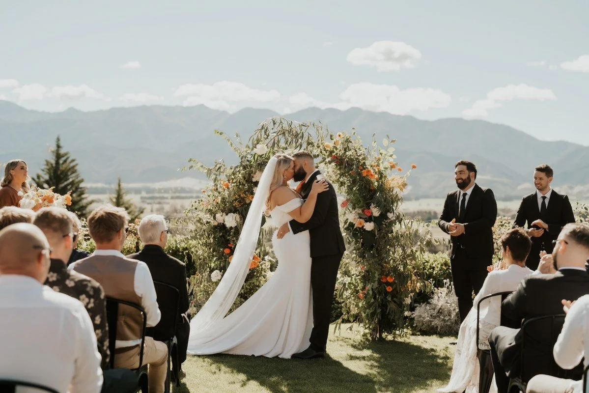 A bride and groom kiss during their outdoor wedding ceremony under a floral arch with mountains in the background, surrounded by guests, on a sunny day.