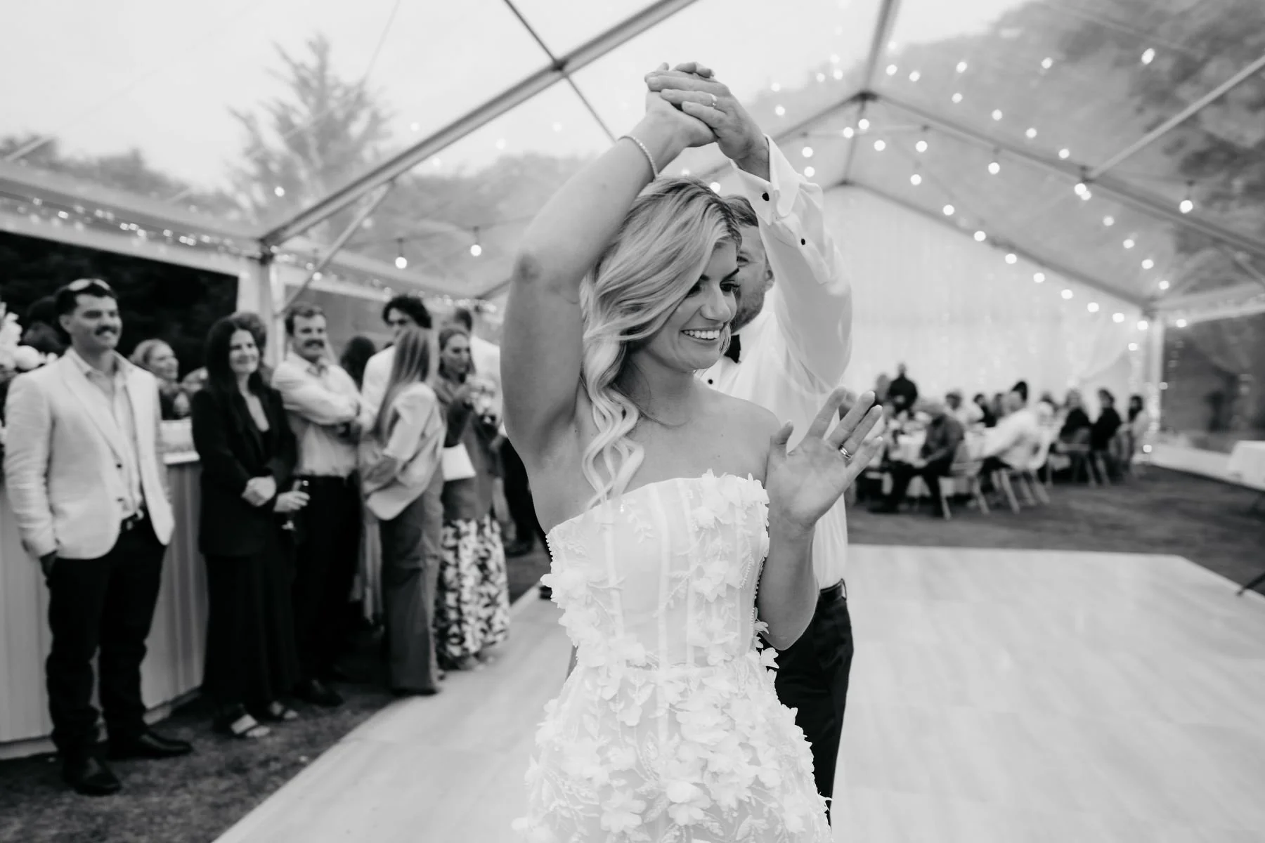 A bride and groom dancing at their wedding reception inside a tent with string lights. The bride is smiling with her eyes closed, wearing a strapless wedding dress with floral details, while the groom, behind her, is holding her hand.