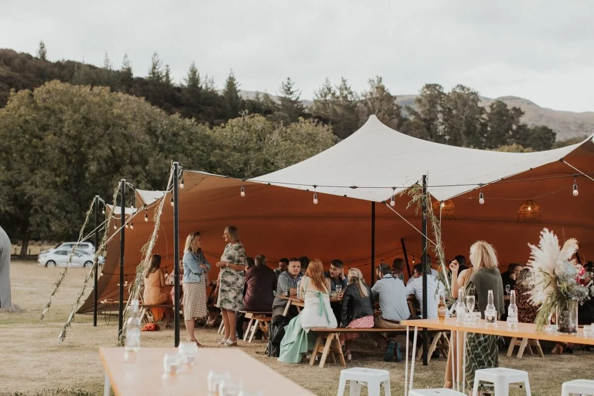 People gathered under a large outdoor tent at a wedding reception or outdoor celebration, with tables, chairs, and string lights, set against a backdrop of trees and hills.