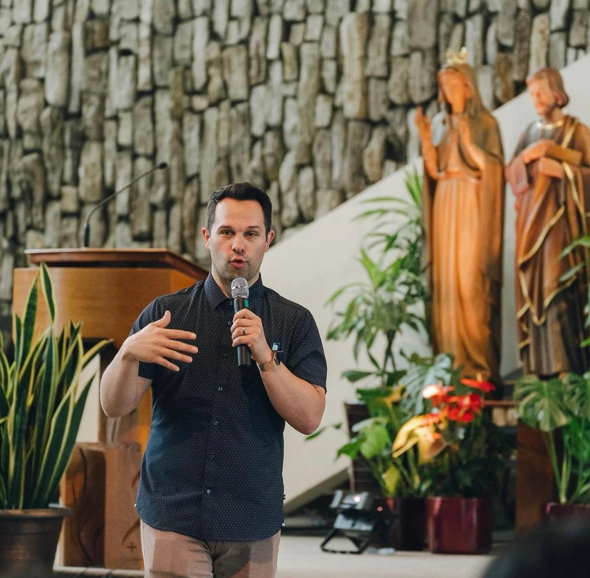 A man speaking into a microphone in a indoor setting with religious statues and plants in the background.