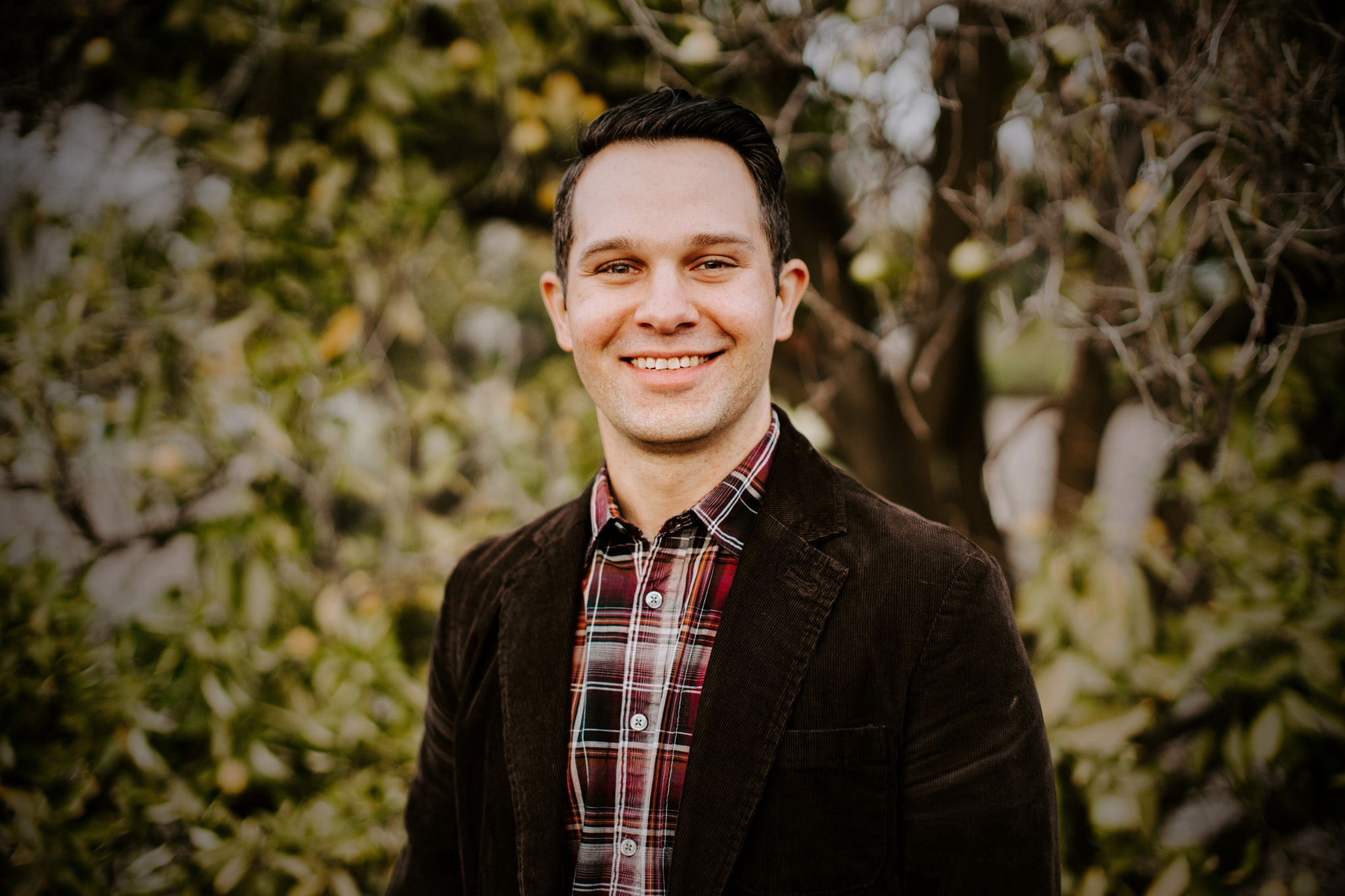 A young man smiling outdoors in front of trees, wearing a dark brown blazer and plaid shirt.