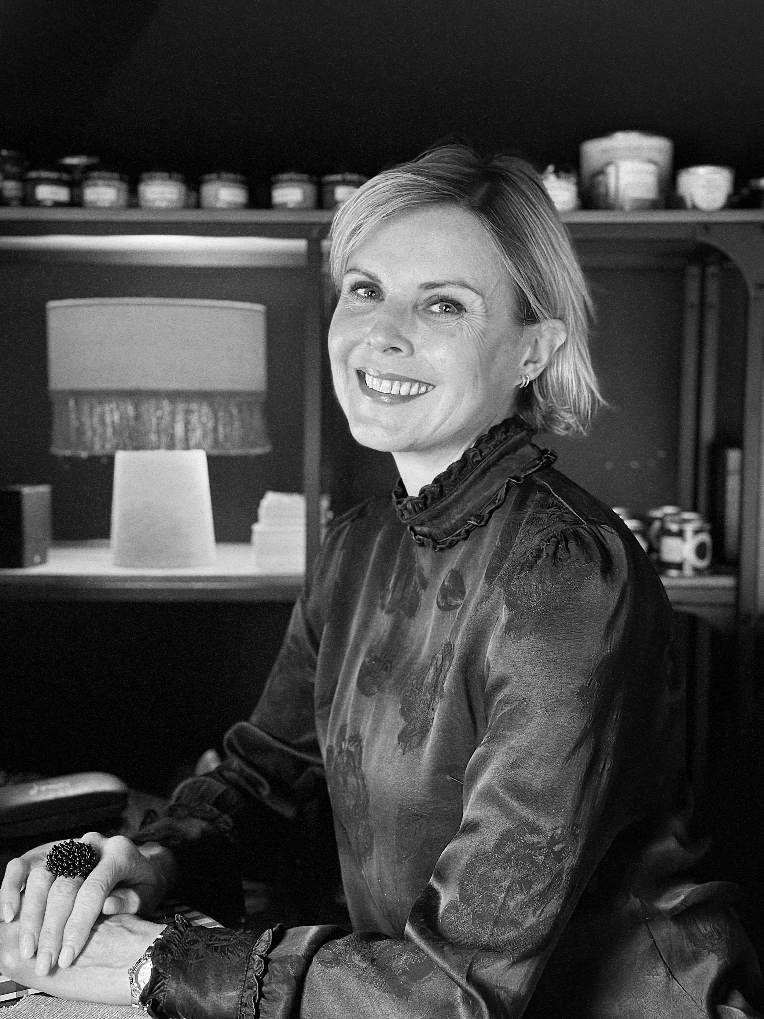 A smiling woman with short hair sitting at a desk in a room with bookshelves and decorative items, in black and white.